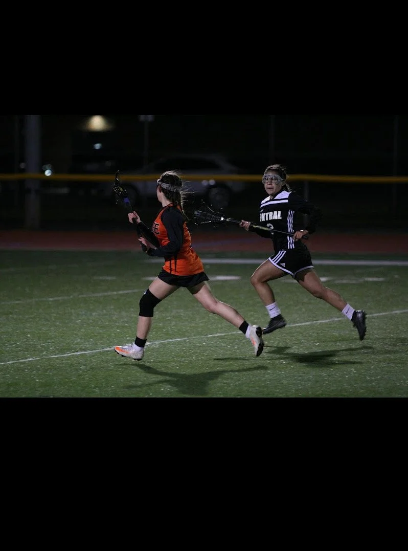 Two young girls playing lacrosse on a field at night, wearing protective goggles and sports jerseys, running after the ball.