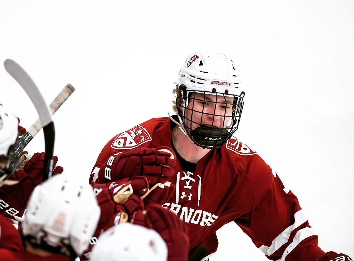 A young male ice hockey player in a red jersey and white helmet smiling and celebrating with teammates on the ice.