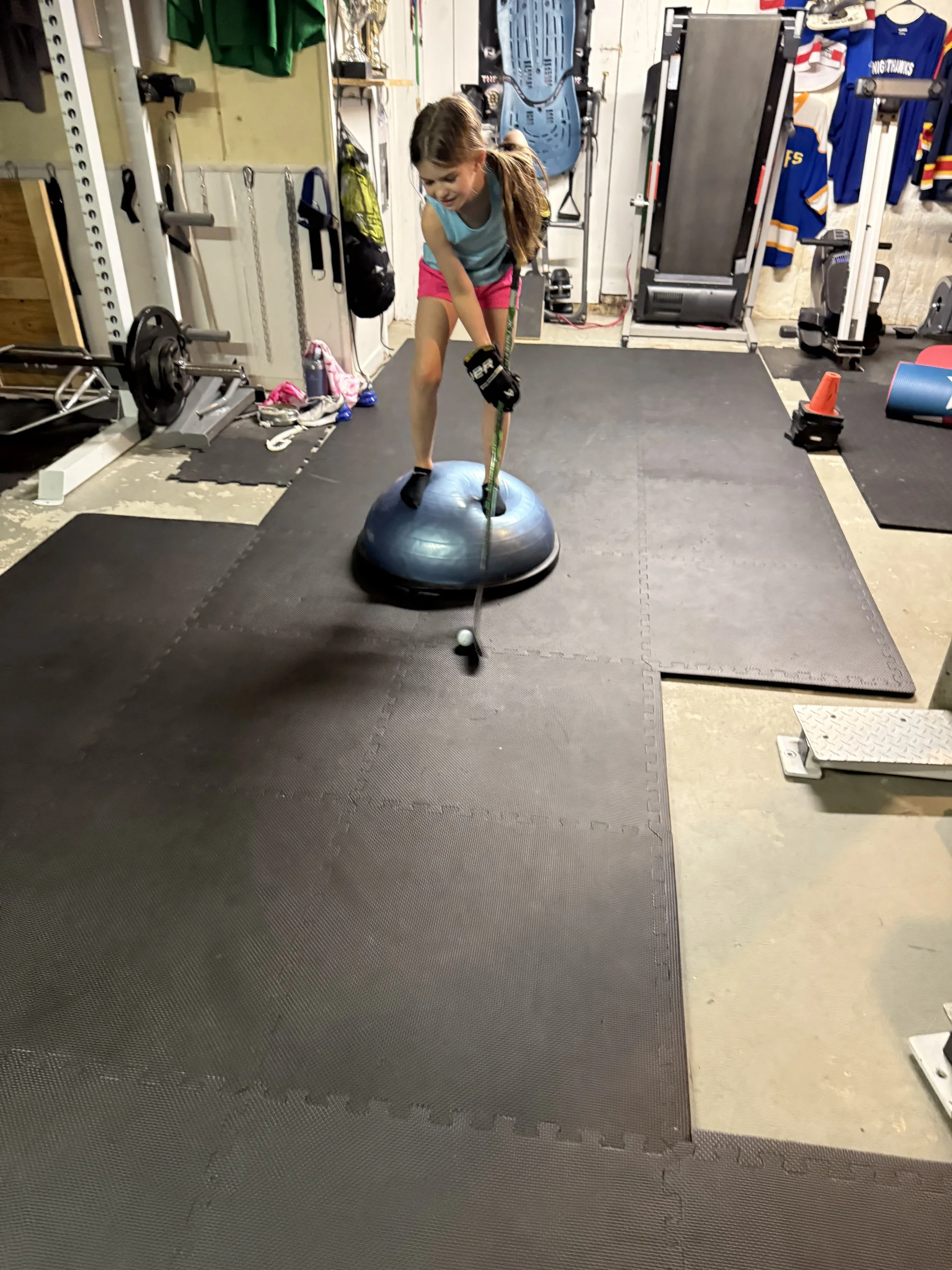 A young girl in a gym or home workout space is standing on a balance wheel, holding a hockey stick, with a golf ball positioned at the end of the stick. She is wearing a turquoise tank top, pink shorts, and hockey gloves, and has brown hair tied back