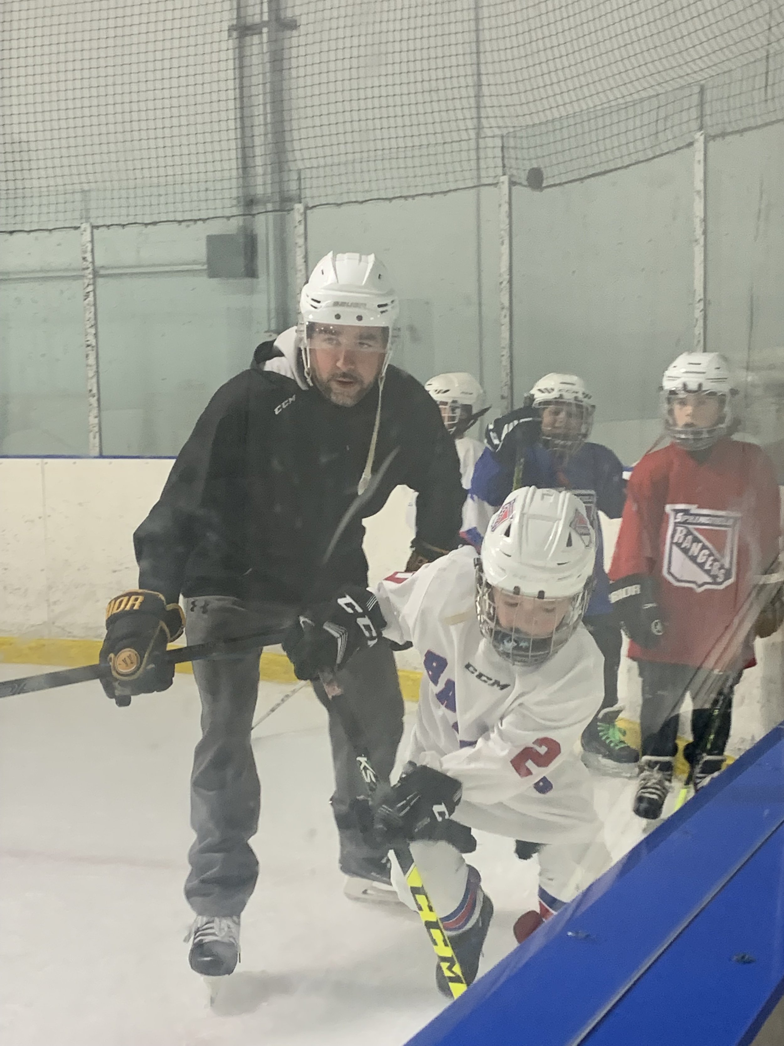 Hockey players on the ice during a game or practice, with players wearing helmets and hockey gear, seen through a glass barrier.
