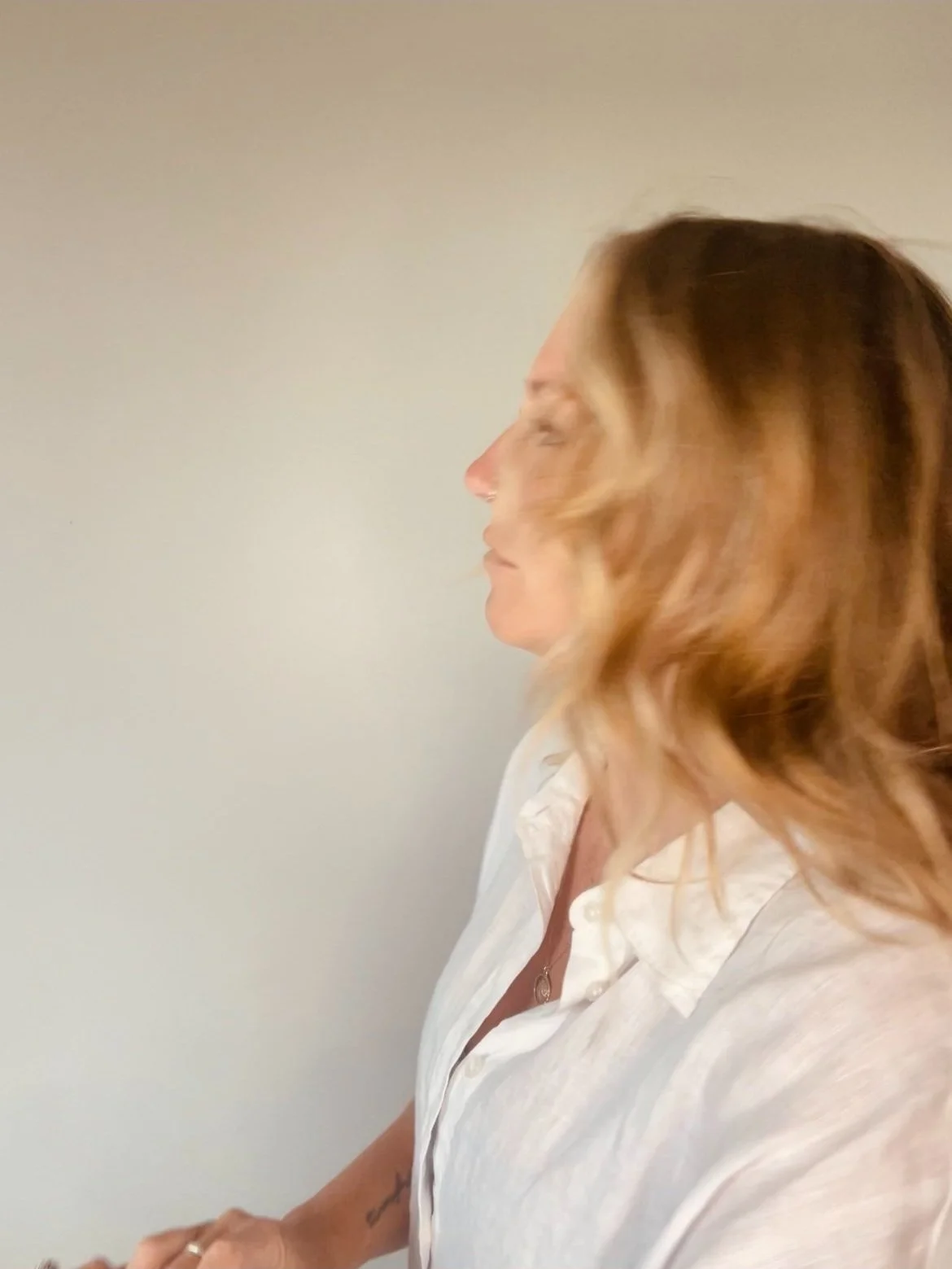 Side profile of a woman with wavy, shoulder-length blonde hair, wearing a white shirt with an open collar, standing against a plain light-colored wall.