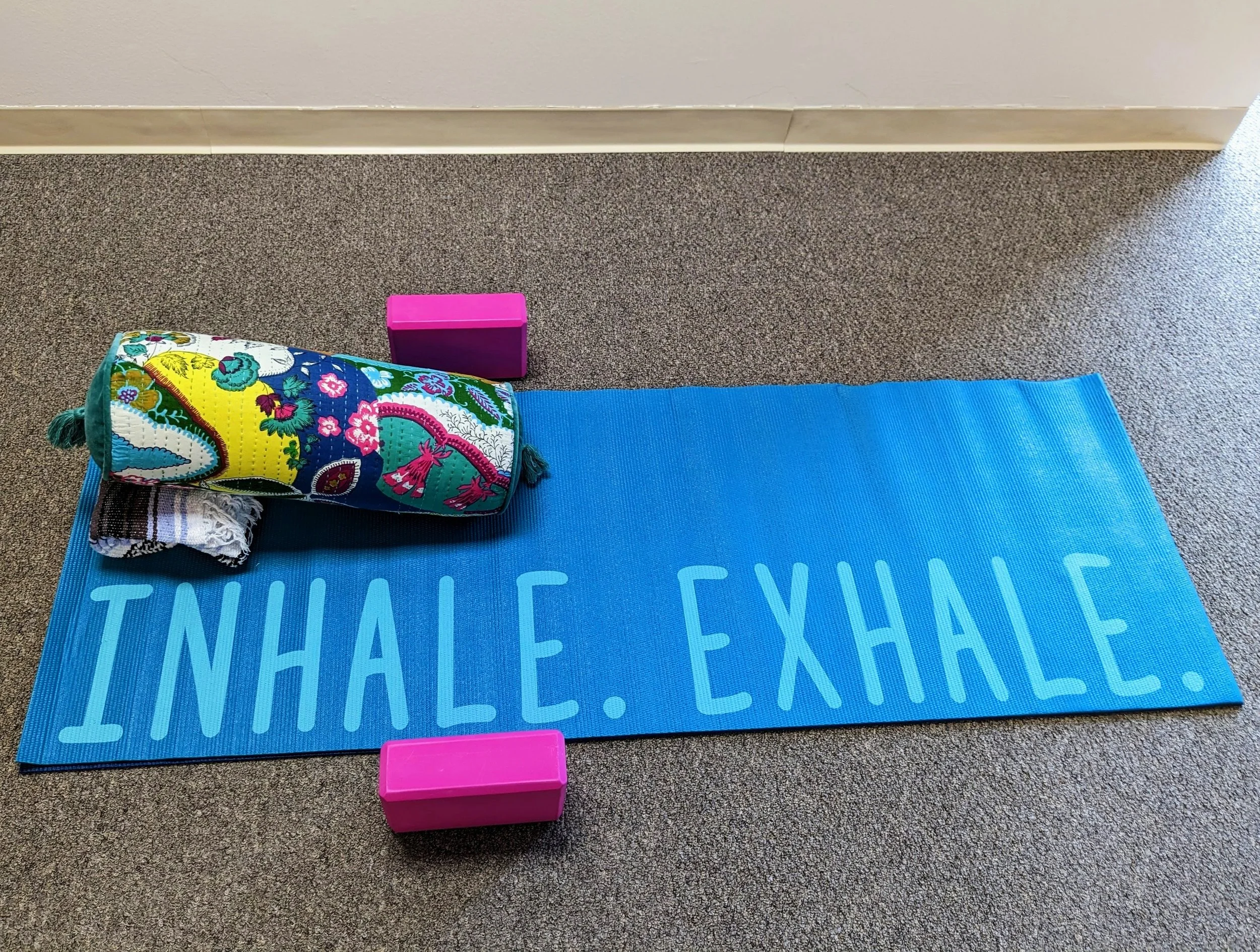 Blue yoga mat with 'INHALE. EXHALE.' text, colorful bolster, striped blanket, and two pink yoga blocks on carpeted floor.