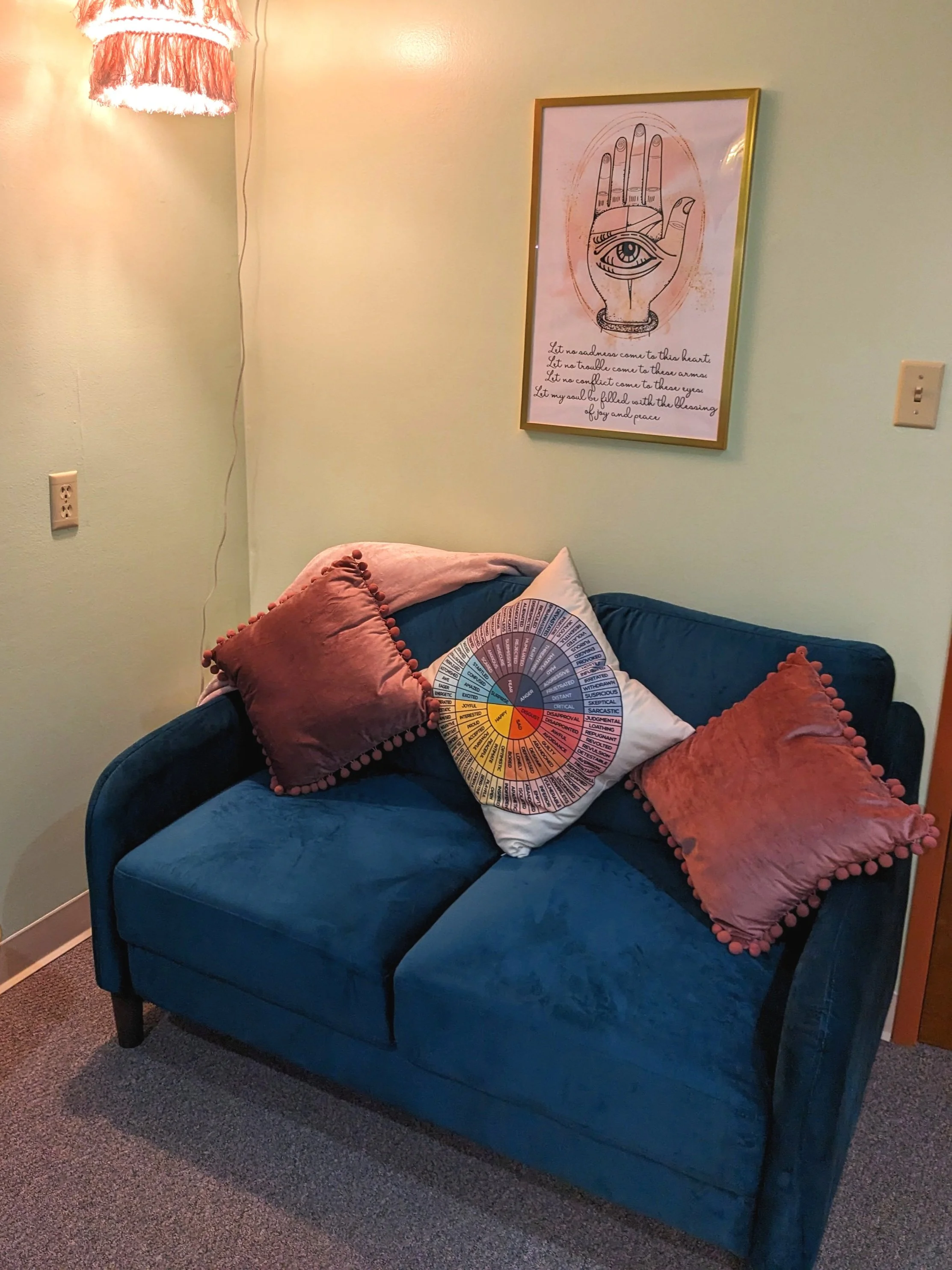 A cozy room with a blue velvet loveseat featuring two pink throw pillows with pom-poms and a multicolored circle design pillow. Above the loveseat, there's a framed poster of a hand with an eye and text underneath. The wall is light green, and there's a hanging lampshade with fringe to the left.