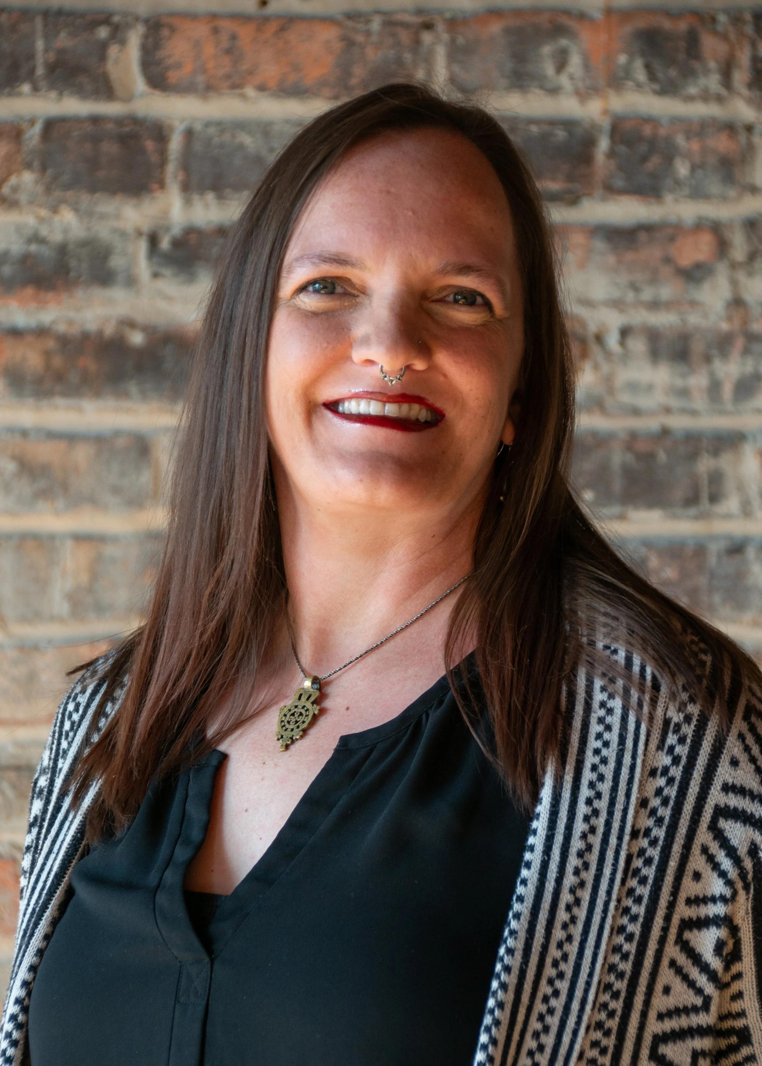 Woman with long brown hair, smiling, wearing a black top and a patterned cardigan, against a brick wall.