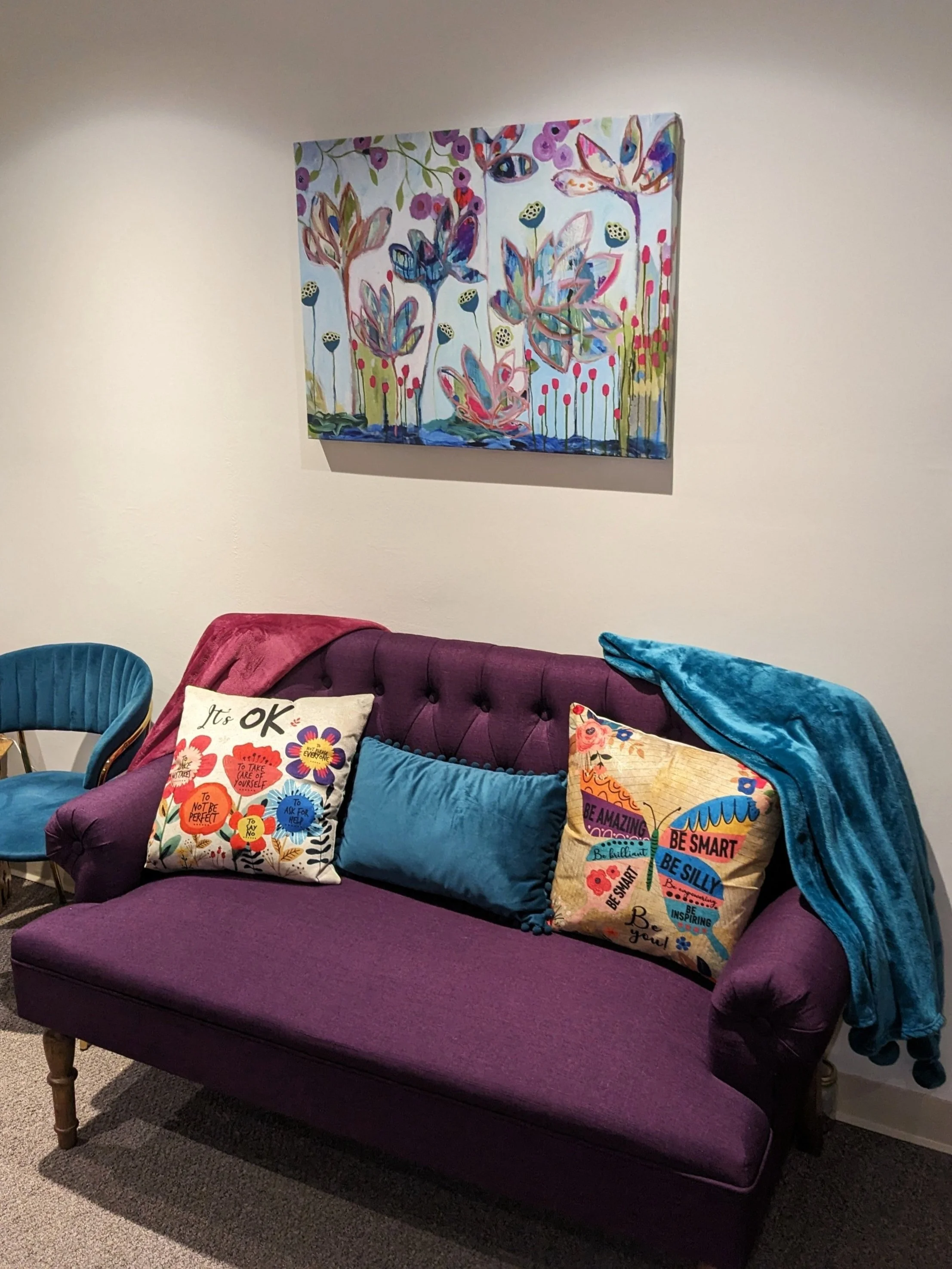 Cozy room with purple sofa, decorative cushions, blue armchair, vibrant abstract flower painting on wall, and teal blanket draped over sofa.