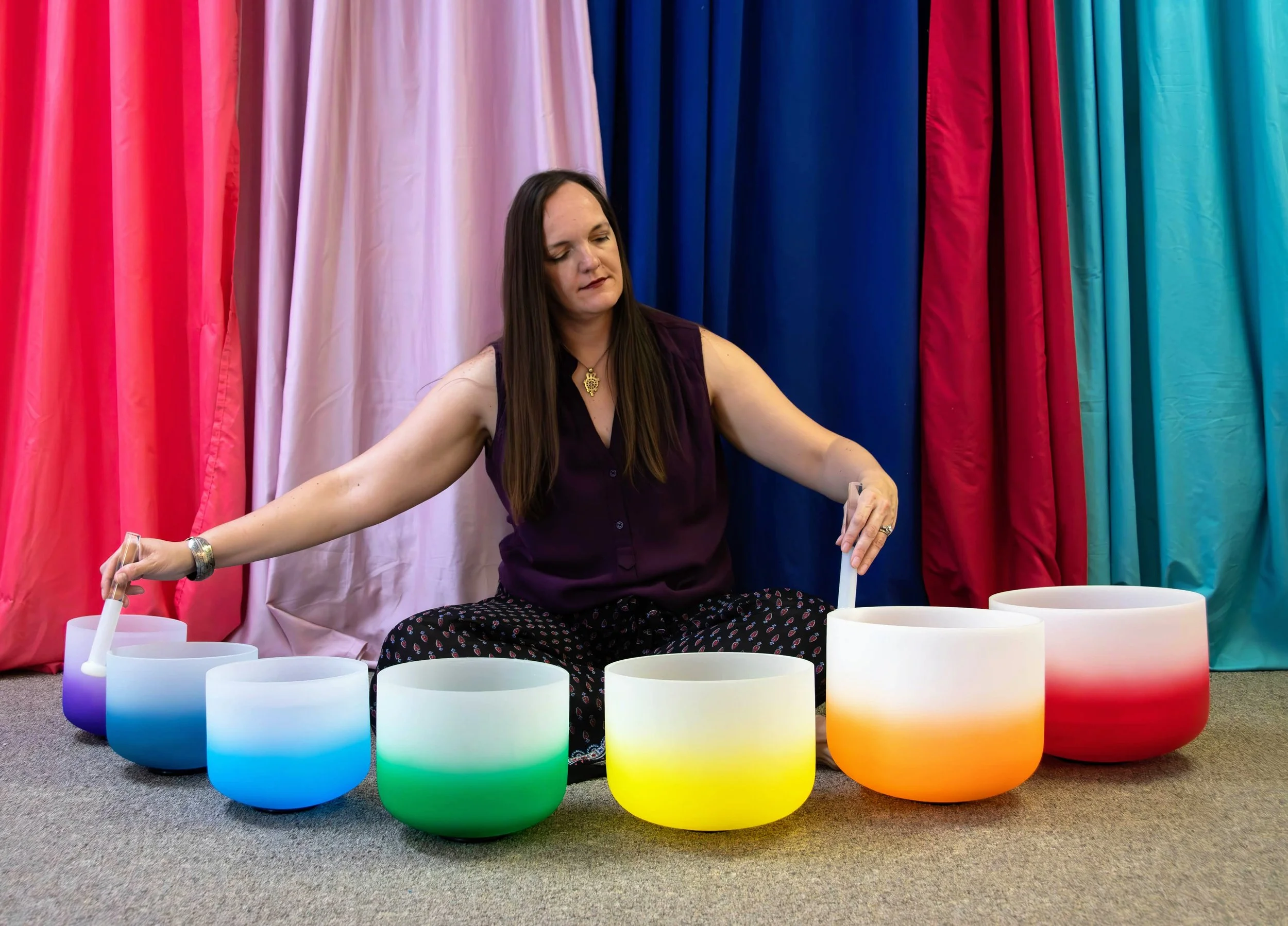 A woman sitting cross-legged on the floor in front of colorful curtains, playing a set of seven rainbow colored crystal singing bowls arranged in a slight semi circle.