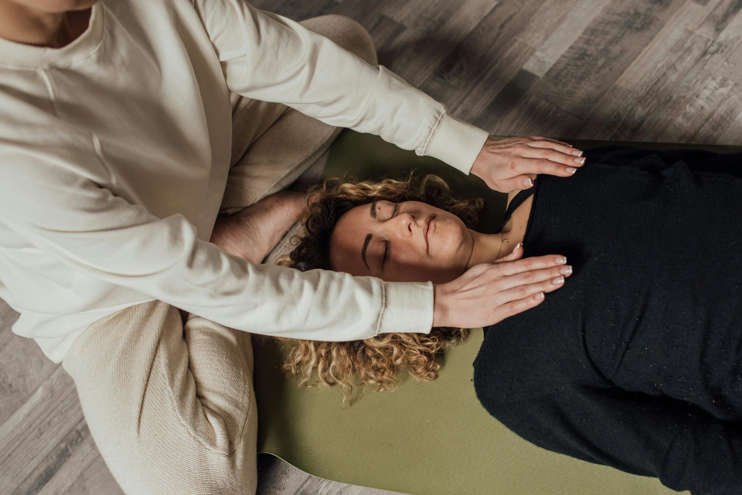 A woman lying on a meditation cushion with her eyes closed, while a person in a cream-colored sweater gently hovers her hands over the person lying down's collar bones for reiki.