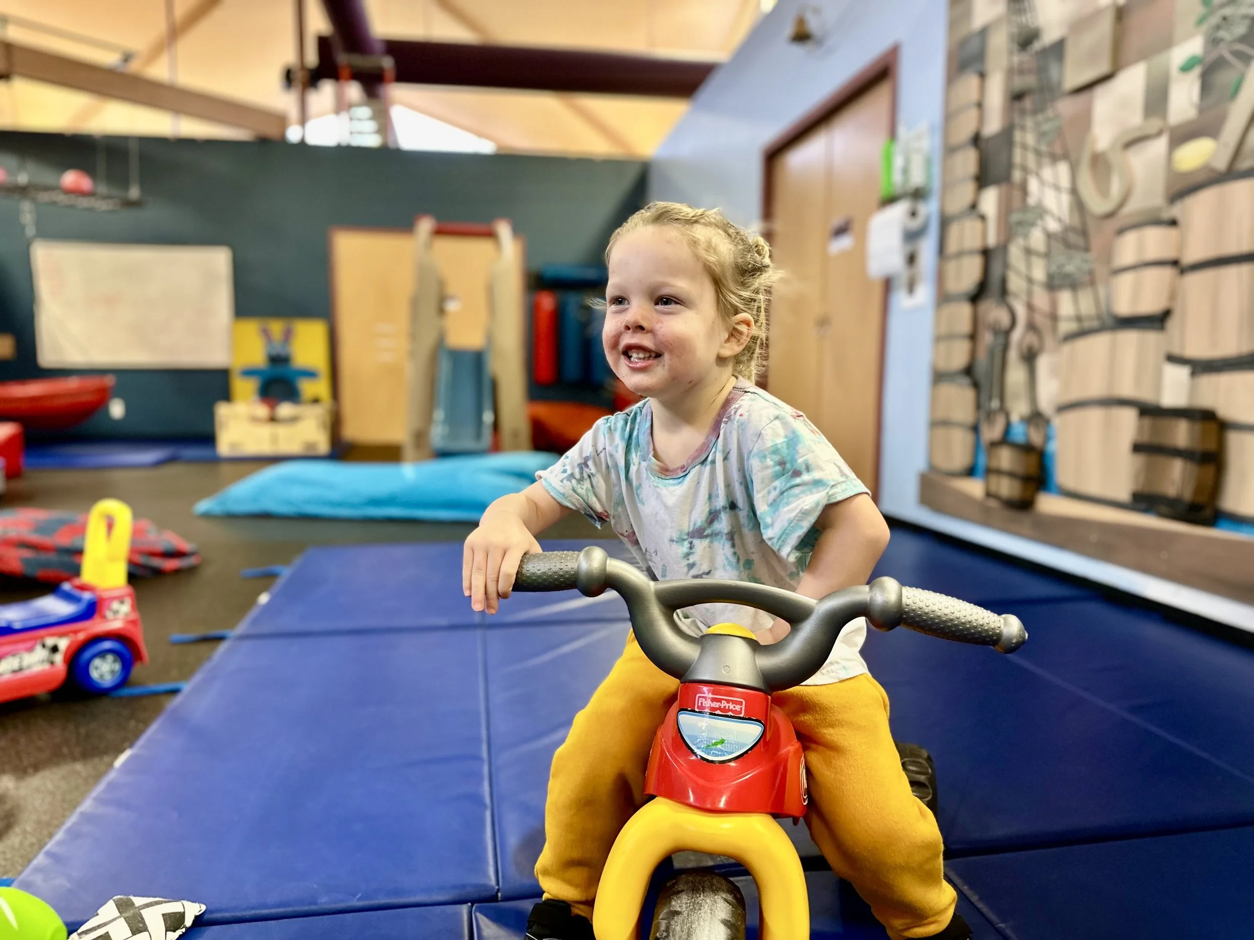 Child riding a toy tricycle indoors on blue mats, with cheerful expression, wearing colorful shirt and yellow pants, in a playroom setting.