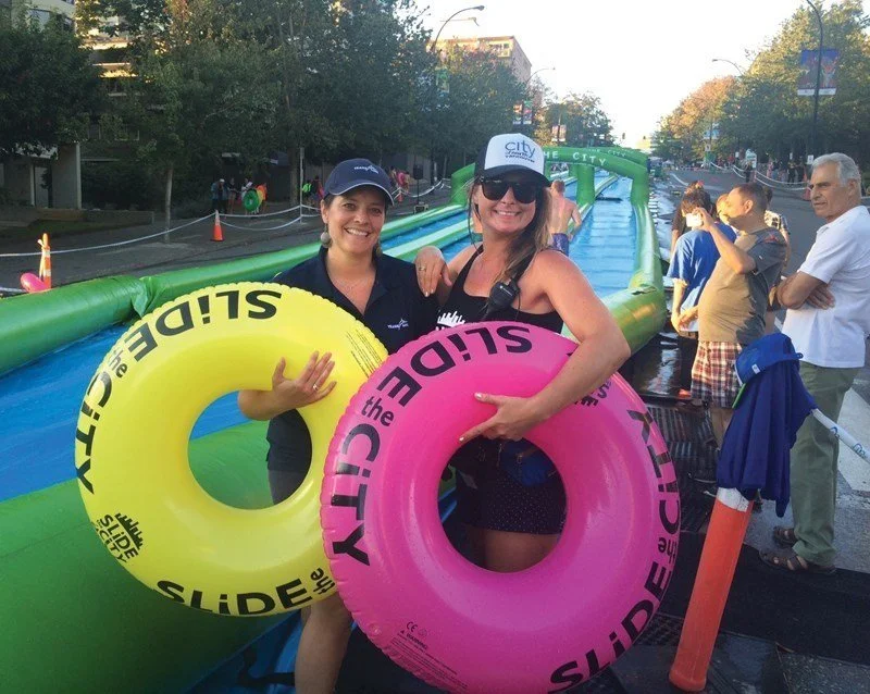 Two women holding inflatable tubes with "Slide the City" branding, standing near an outdoor water slide on a city street, surrounded by other people and trees.