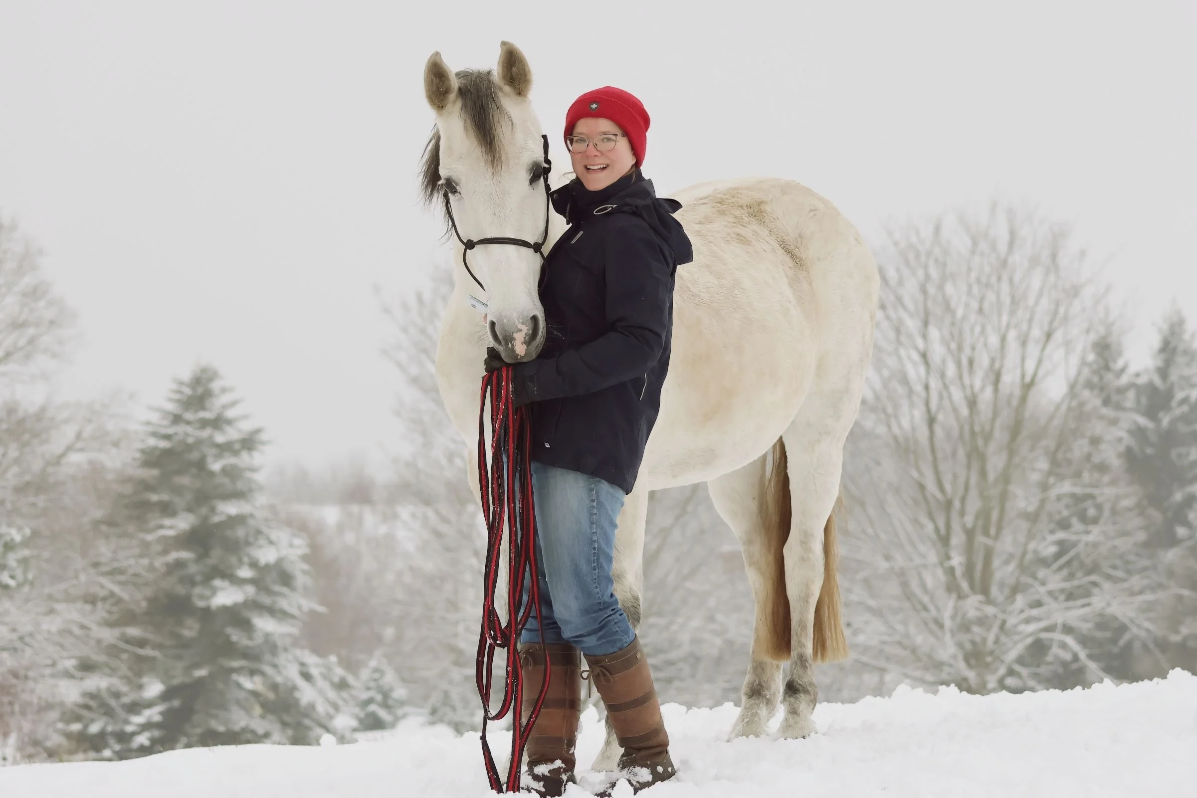 Eine Frau in Winterkleidung steht im Schnee, hält die Zügel eines weißen Pferdes und lächelt in die Kamera. Im Hintergrund sind schneebedeckte Bäume.