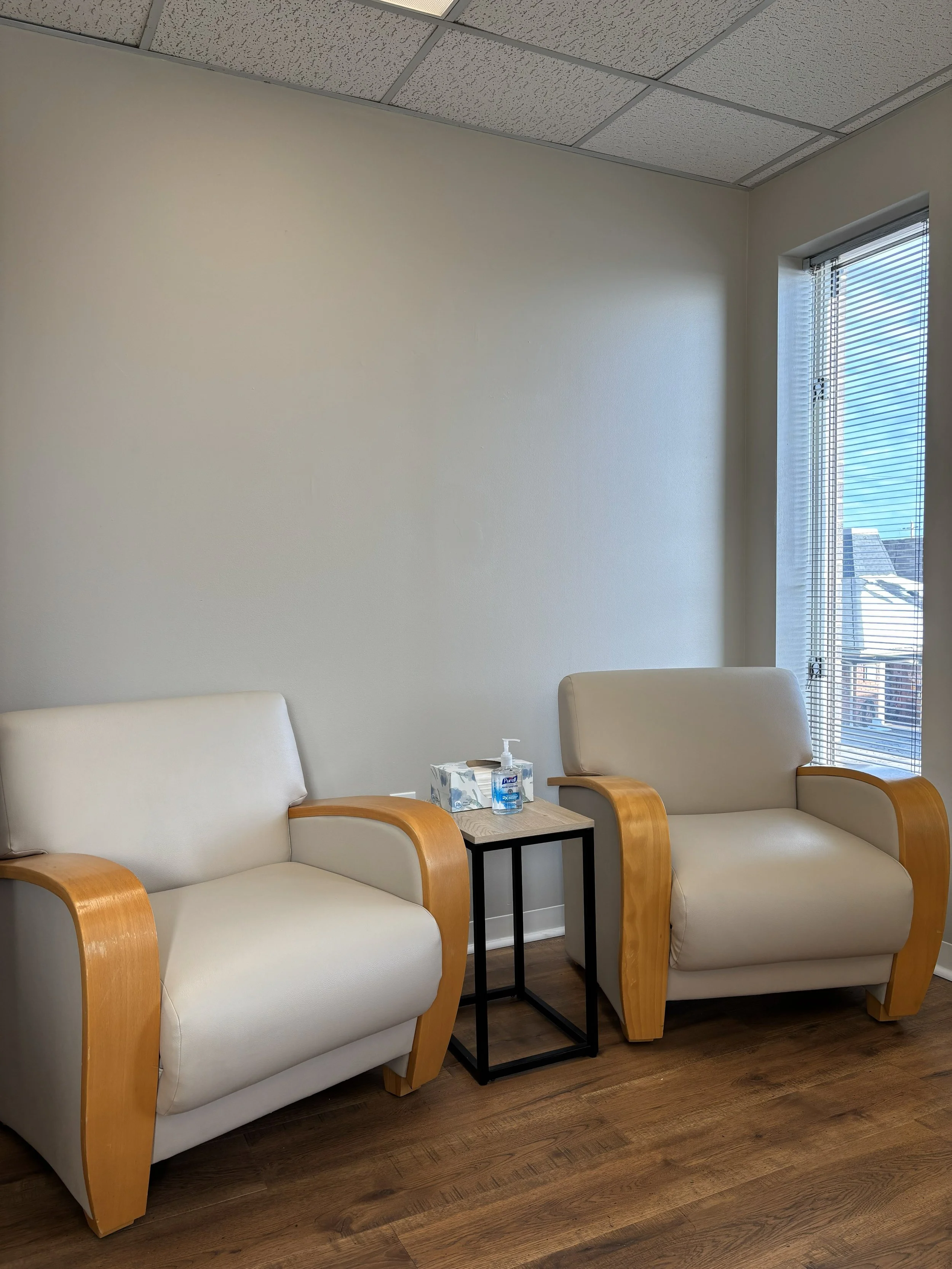 Two beige armchairs with wooden arms are placed beside each other in a waiting room. A small black side table with tissues and hand sanitizer is between them. A window with blinds is on the right, revealing a building outside. The room has a wood floor and a ceiling with tile panels.