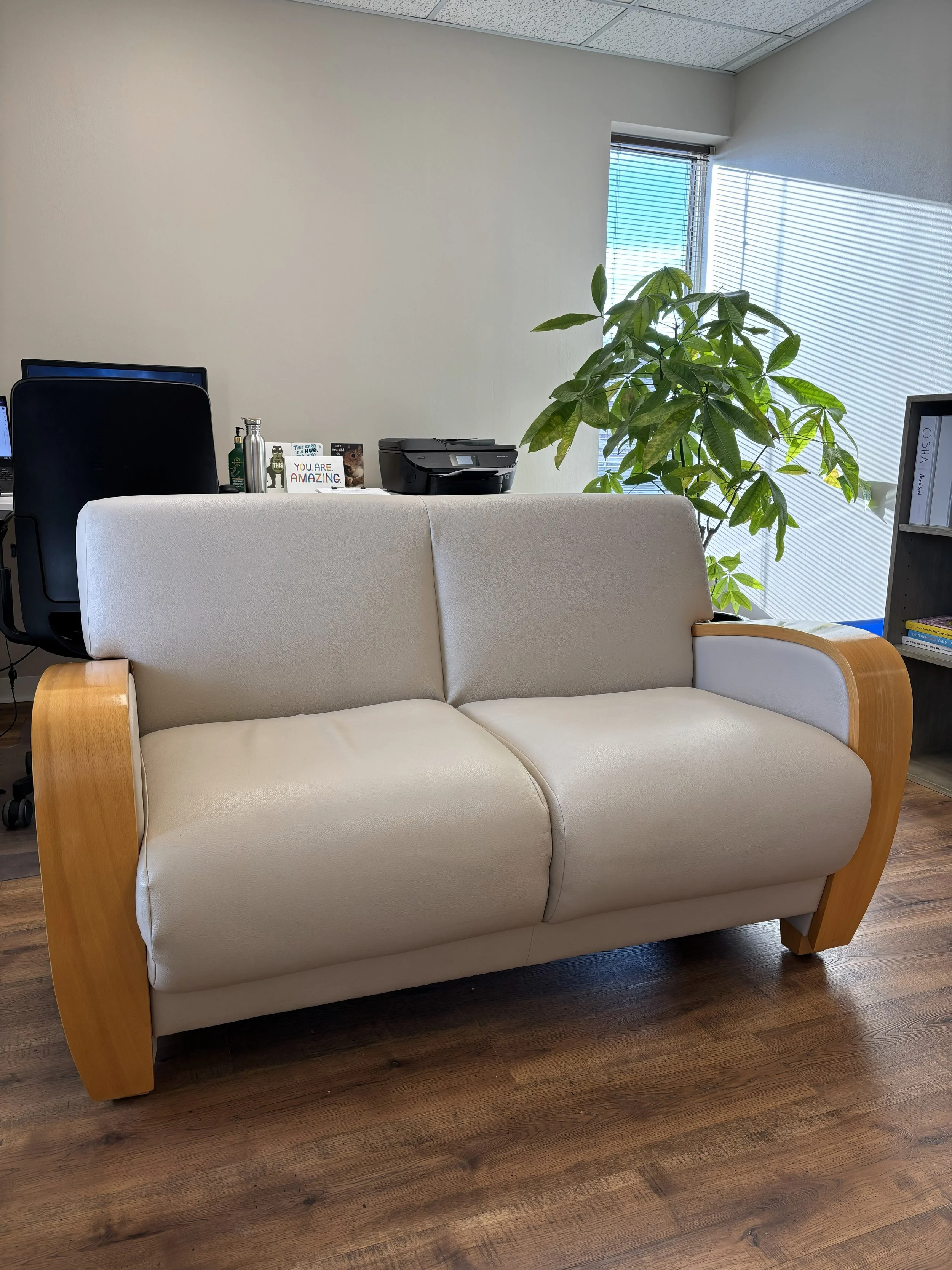 An office sofa with beige upholstery and wooden armrests, situated on a wooden floor near a window with blinds, with a large green leafy plant in the background.