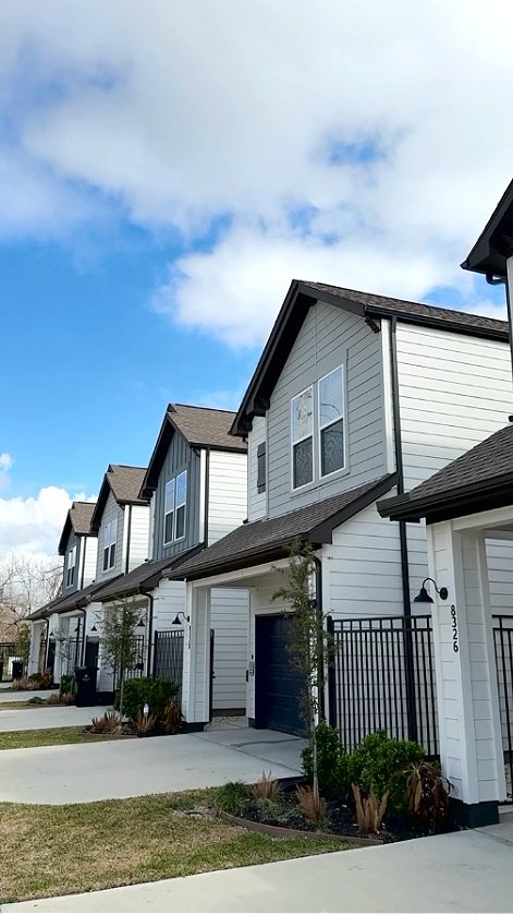 Row of modern suburban townhouses with white siding and dark roofs, featuring small front gardens, driveway, and clear blue sky in the background.
