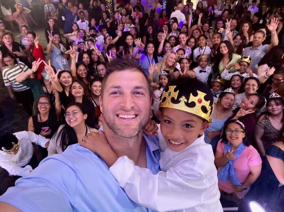 Group photo of a man with a child wearing a crown, surrounded by a large, joyful crowd. People are smiling, posing, and some are wearing crowns or tiaras. The setting appears festive.