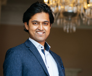 Man with dark hair smiling in a blue suit and white shirt indoors with a chandelier in the background.