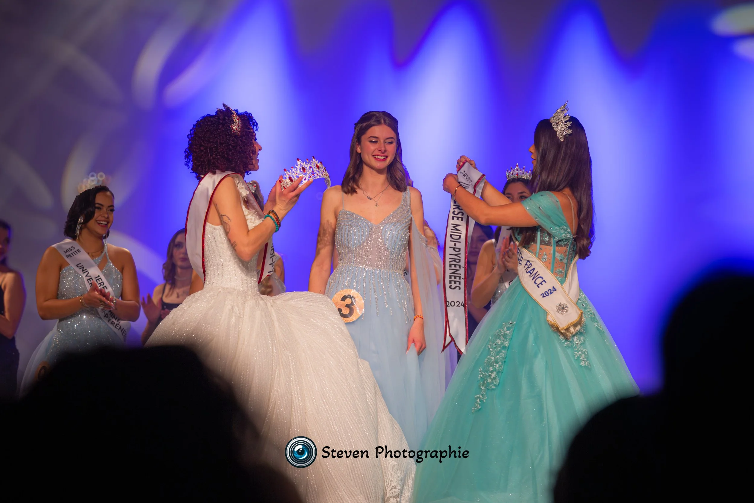 Concours de beauté avec des participantes en robes de soirée, l'une reçoit une couronne et une écharpe gagnante, scène éclairée en violet.