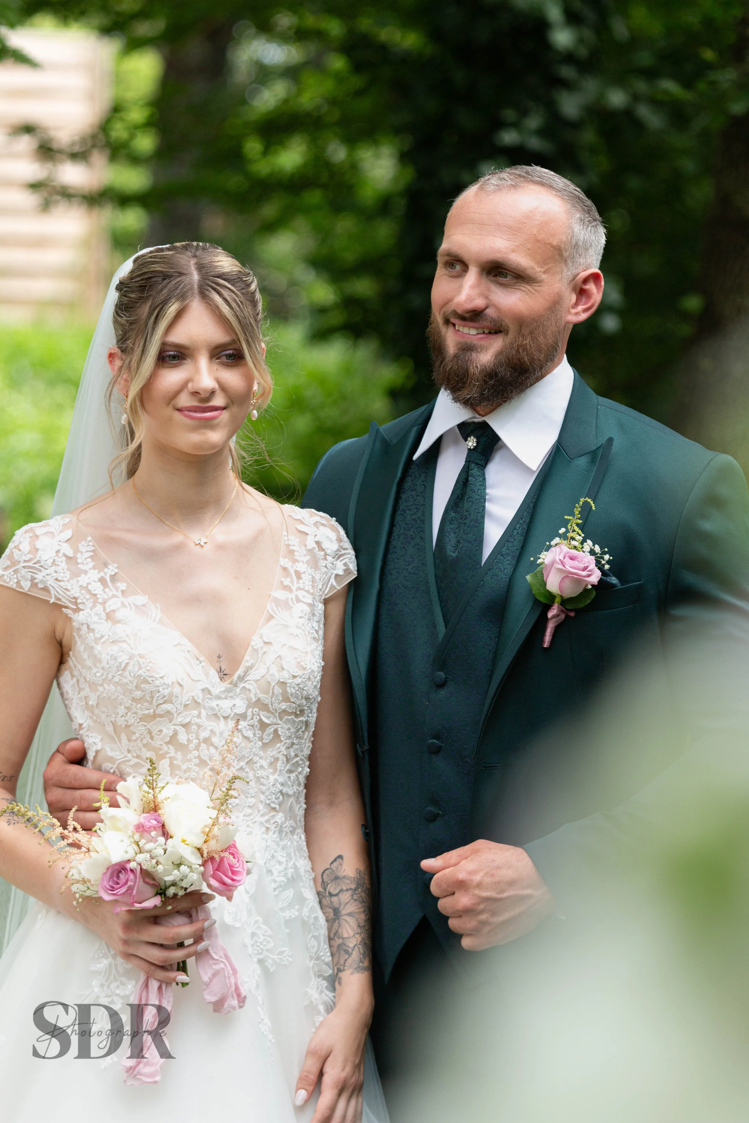 Un couple de mariés pose pour une photo en extérieur, la mariée porte une robe blanche avec des dentelles et tient un bouquet de fleurs, le marié est vêtu d'un costume vert avec une cravate.