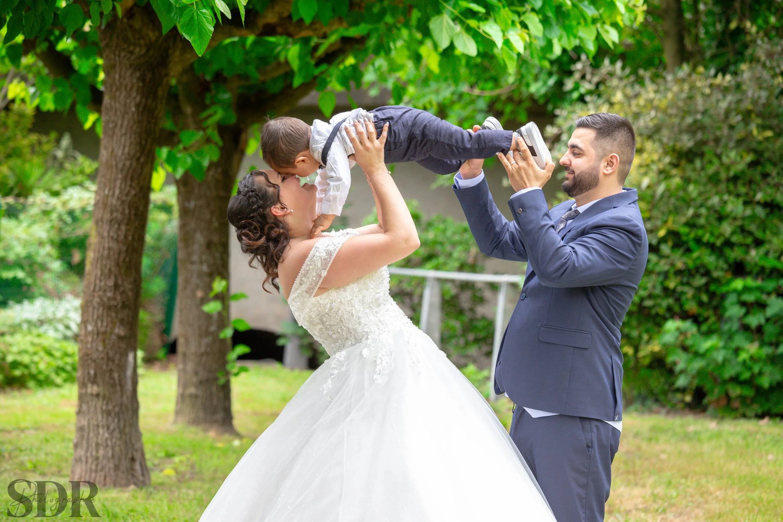Un couple élégant, la femme en robe de mariée et l'homme en costume, soulève un enfant en l'air dans un jardin verdoyant, montrant un moment de joie familiale.