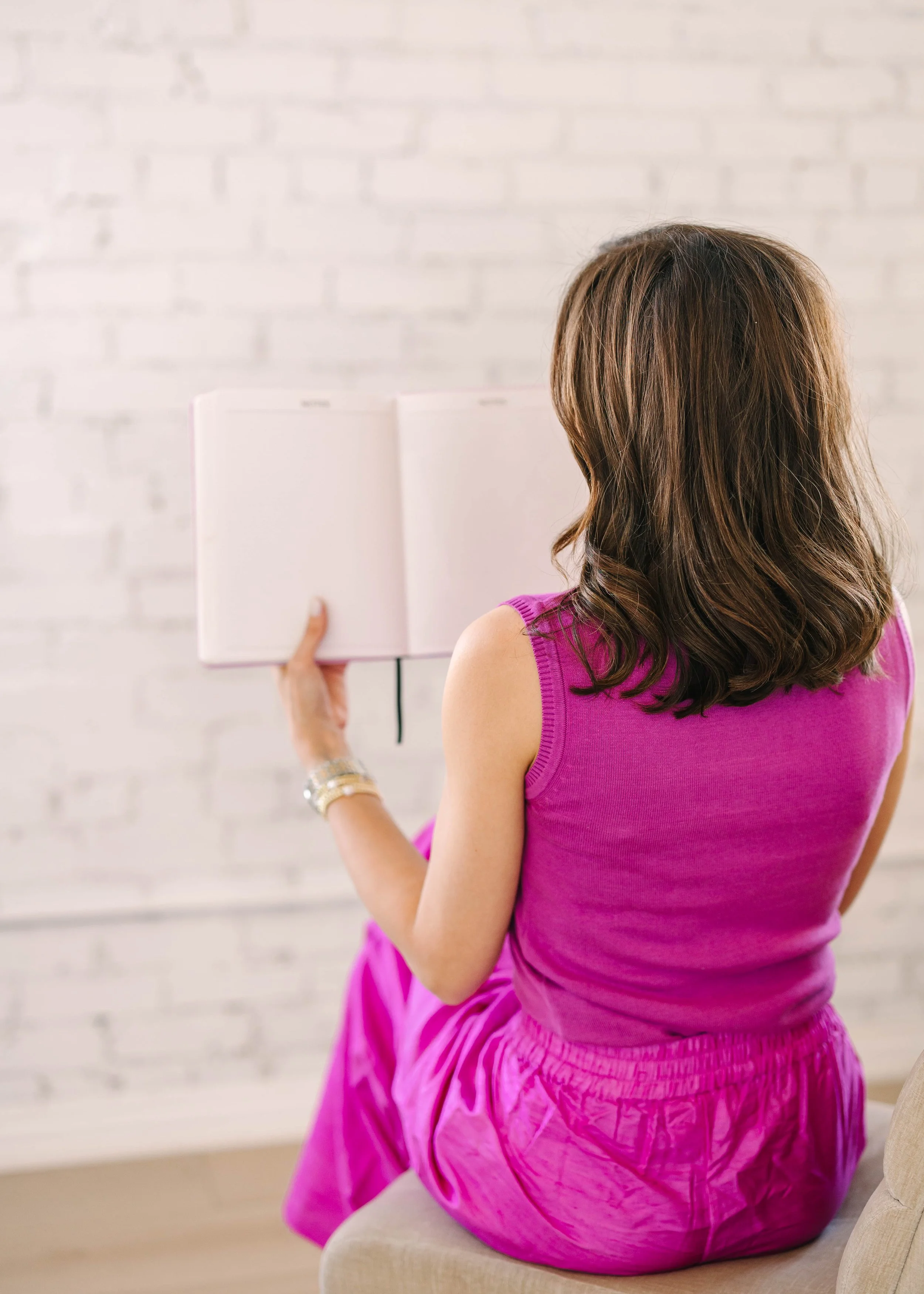 A woman in a pink sleeveless top and pink pants sitting on a beige chair, reading a white open notebook or journal, with a white brick wall in the background.