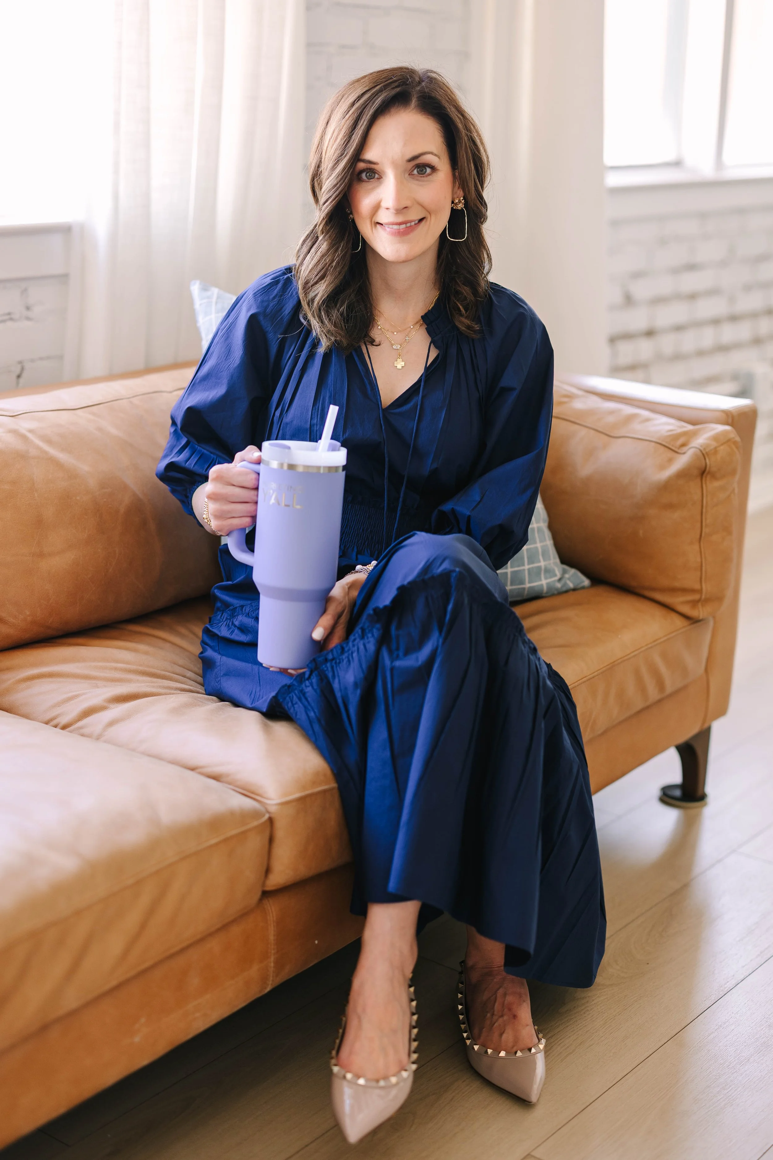 A woman with shoulder-length brown hair sitting on a tan leather couch, holding a lavender travel mug, wearing a navy blue outfit and beige high heels with studs, in a bright room with white brick walls and large windows.