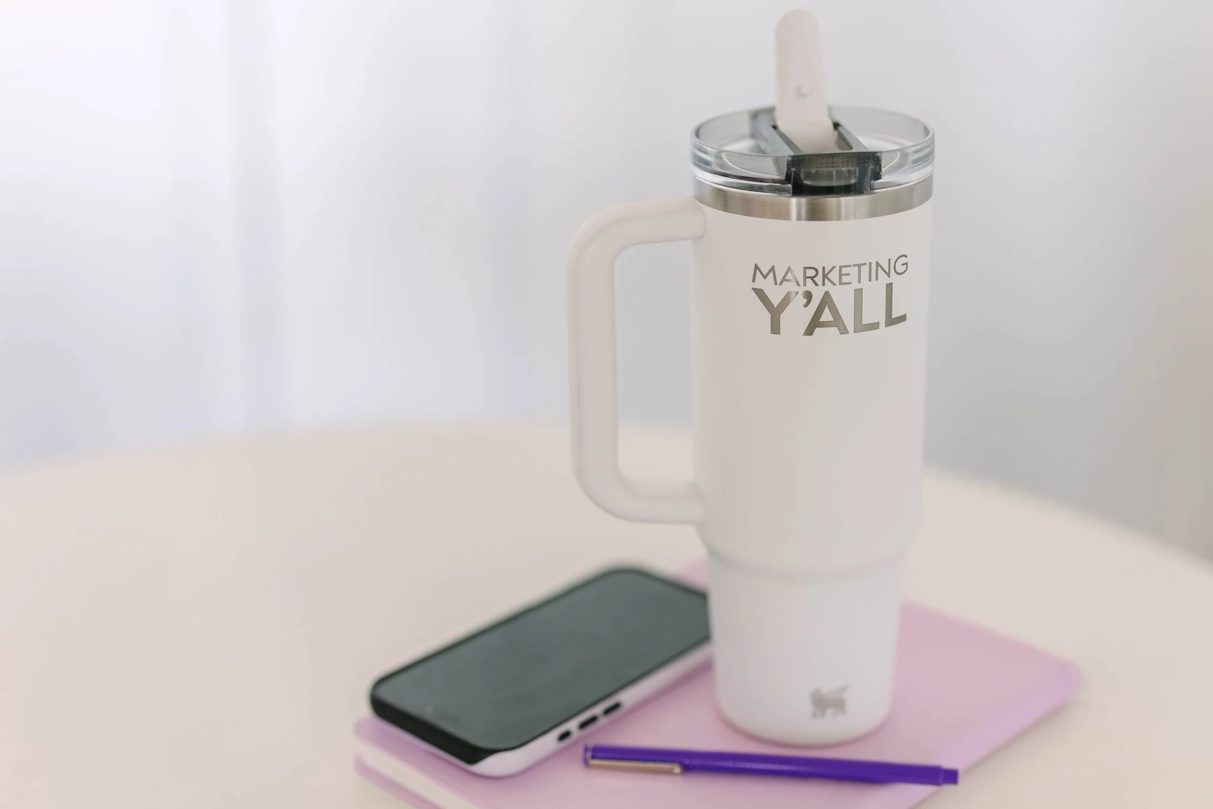 White tumbler with a straw and the words "Marketing Y'all" printed on it, a black smartphone, a purple pen, and a pink notebook on a white table.