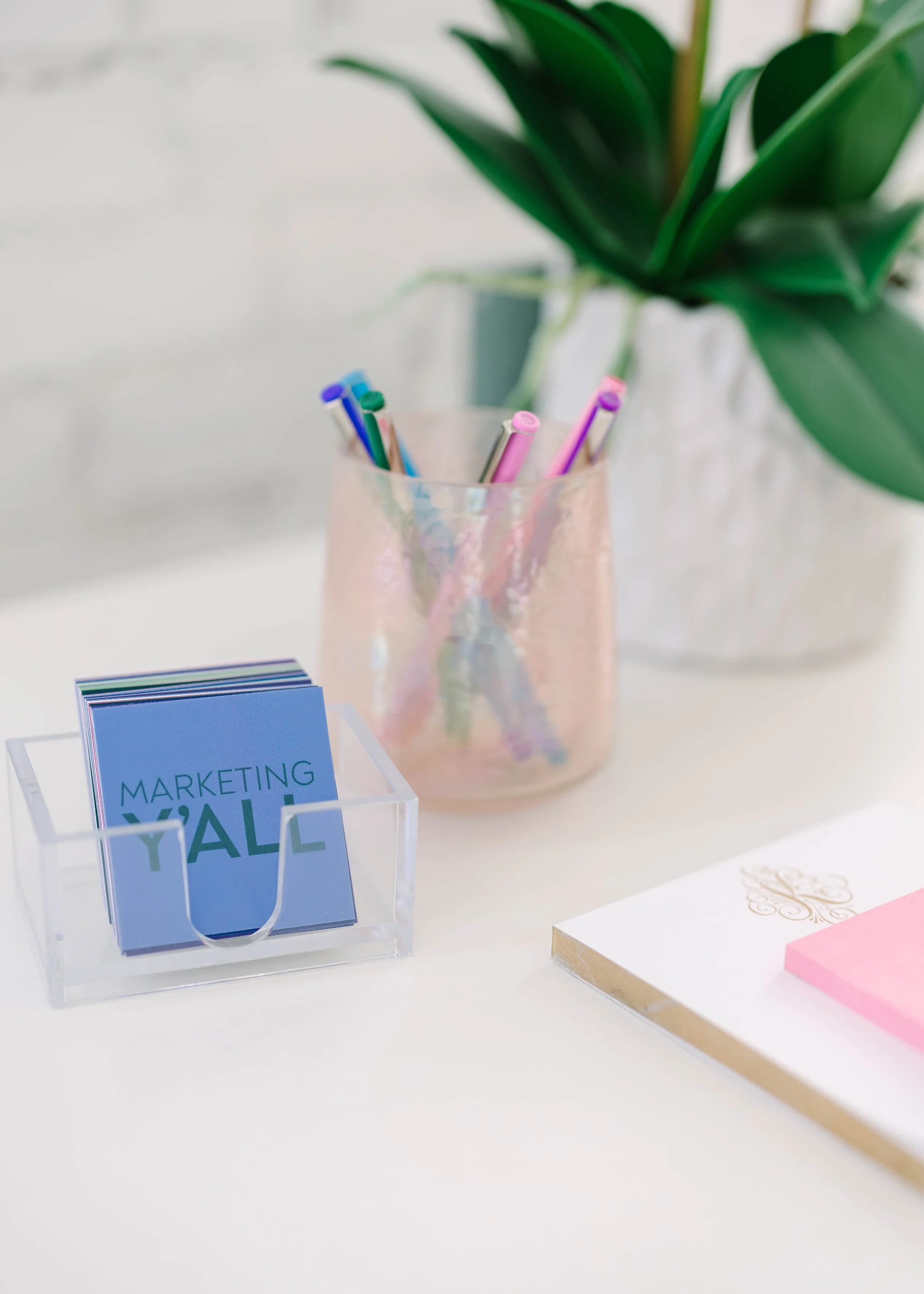 A clear plastic container holding small paper cards with the words "MARKETING YALL" in blue and green. Behind it, a pink transparent cup with colorful pens. To the right, part of a white notepad with gold and pink accents, and a large green plant in a white pot in the background.