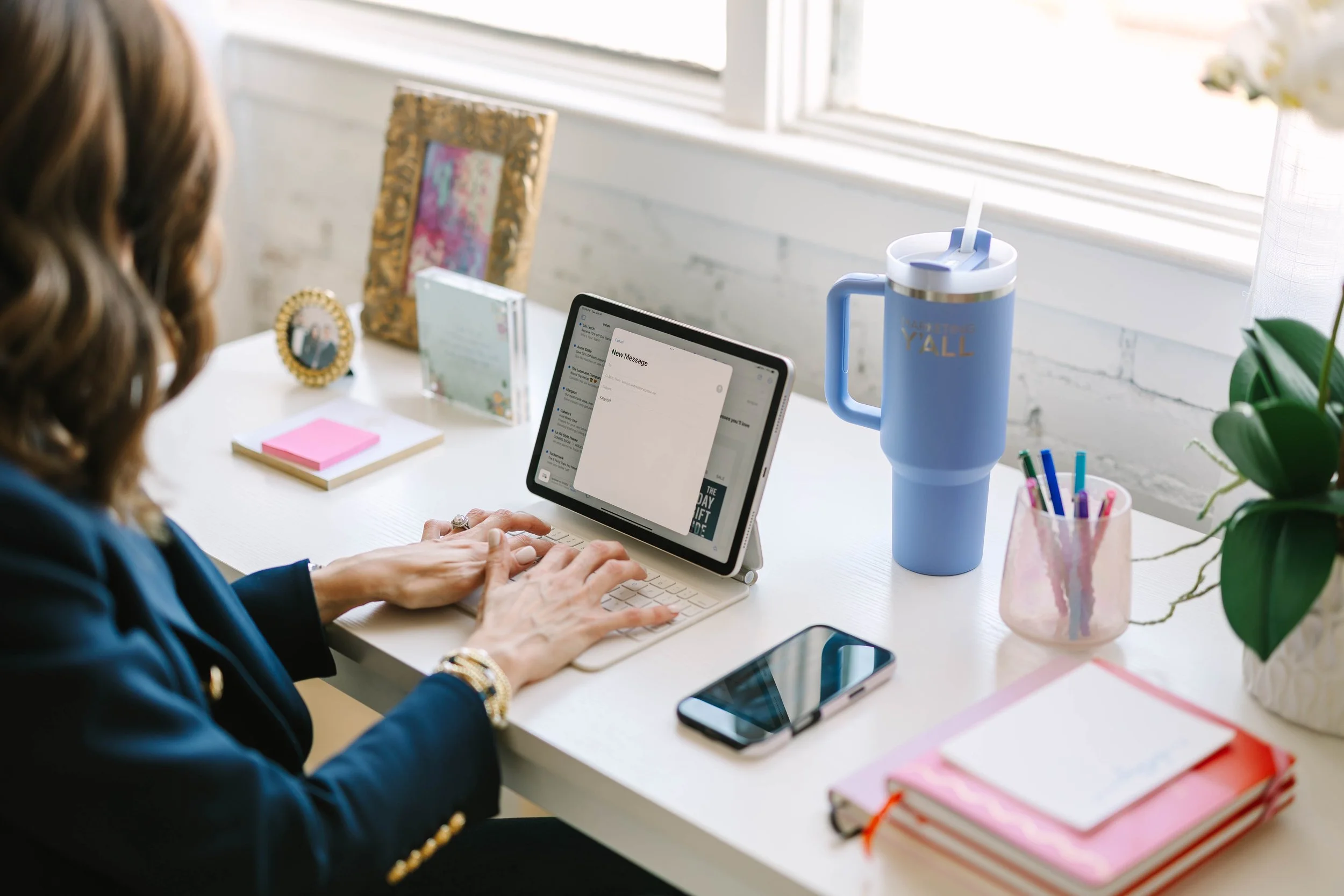 Person using a tablet to type a message at a white desk with various office supplies, including a smartphone, notebooks, a pink cup with pens, a large blue tumbler with a straw, and a plant near a window.