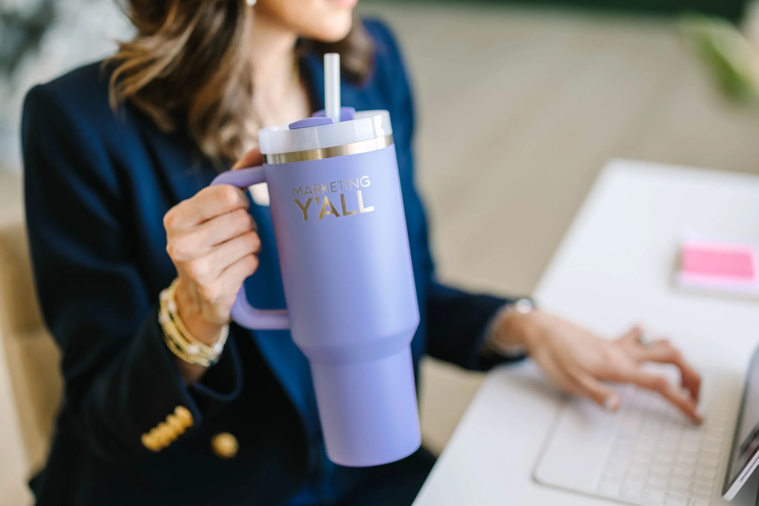 A woman in a dark blazer holding a purple travel mug with gold lettering that reads 'MARKETING Y'ALL'. She is sitting at a desk using a laptop. The background is blurred.