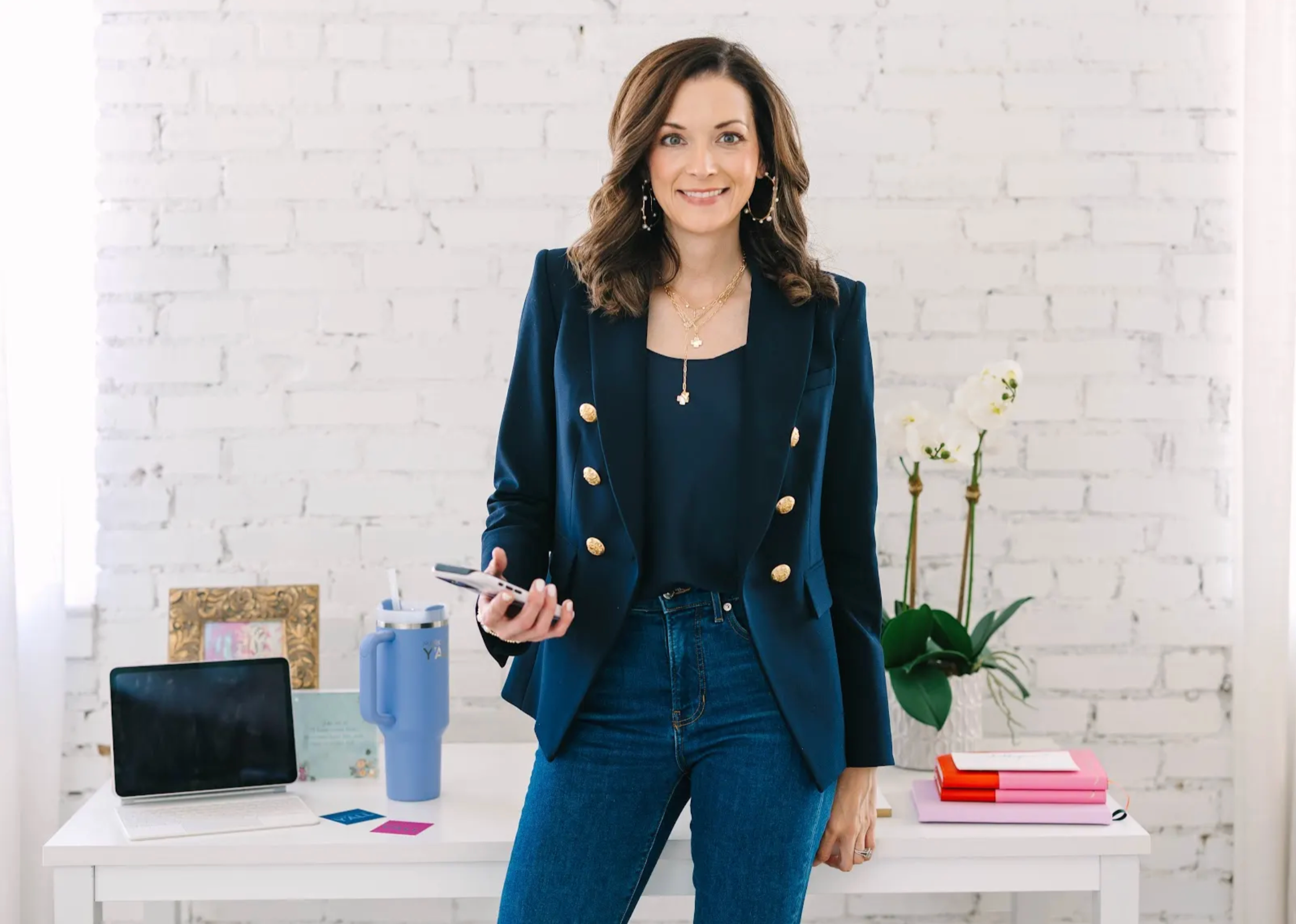 A woman standing in an office, smiling, holding a smartphone in her right hand. She is wearing a navy blazer with gold buttons, a black top, and blue jeans. The office has a white brick wall, a white desk with a laptop, a blue thermos, a framed picture, and colorful folders. There are white orchids in a vase on the desk.