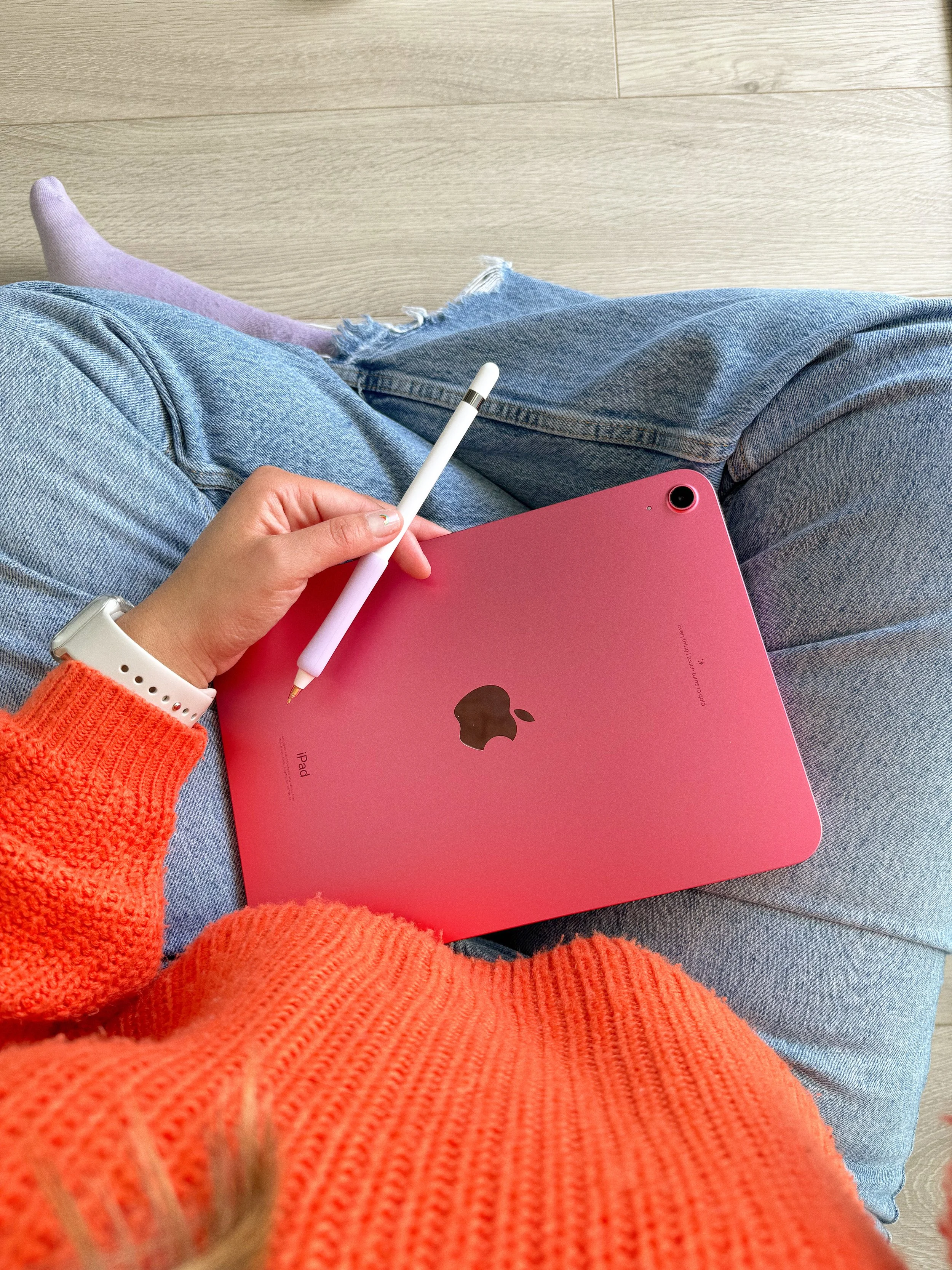 Person sitting cross-legged on a light-colored wooden floor, holding a pink iPad and a white stylus, wearing a bright orange sweater, ripped jeans, and a smartwatch on their left wrist.