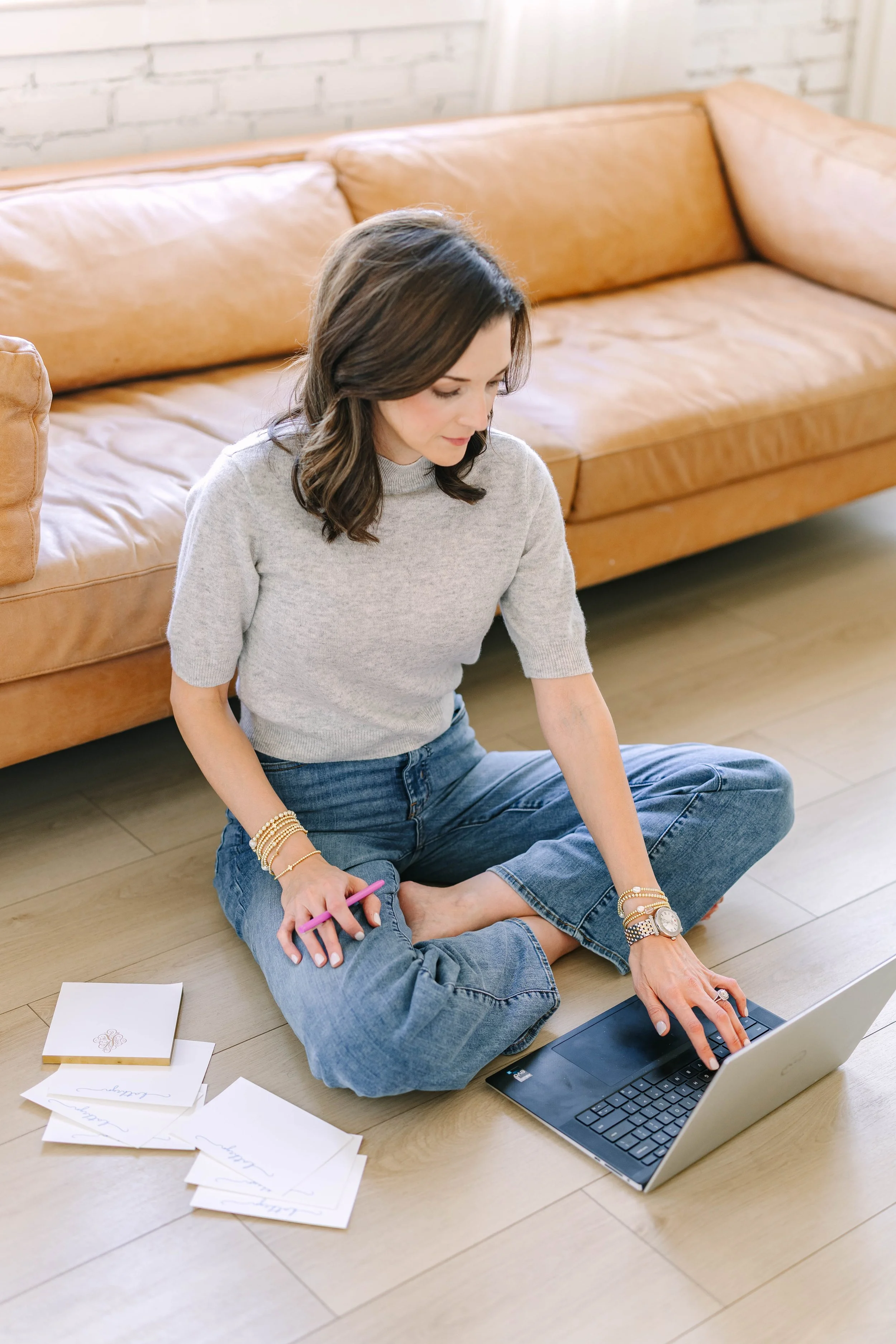 Woman sitting on the floor with a laptop, working or studying, with notes and papers nearby, in a room with a leather sofa and brick wall.