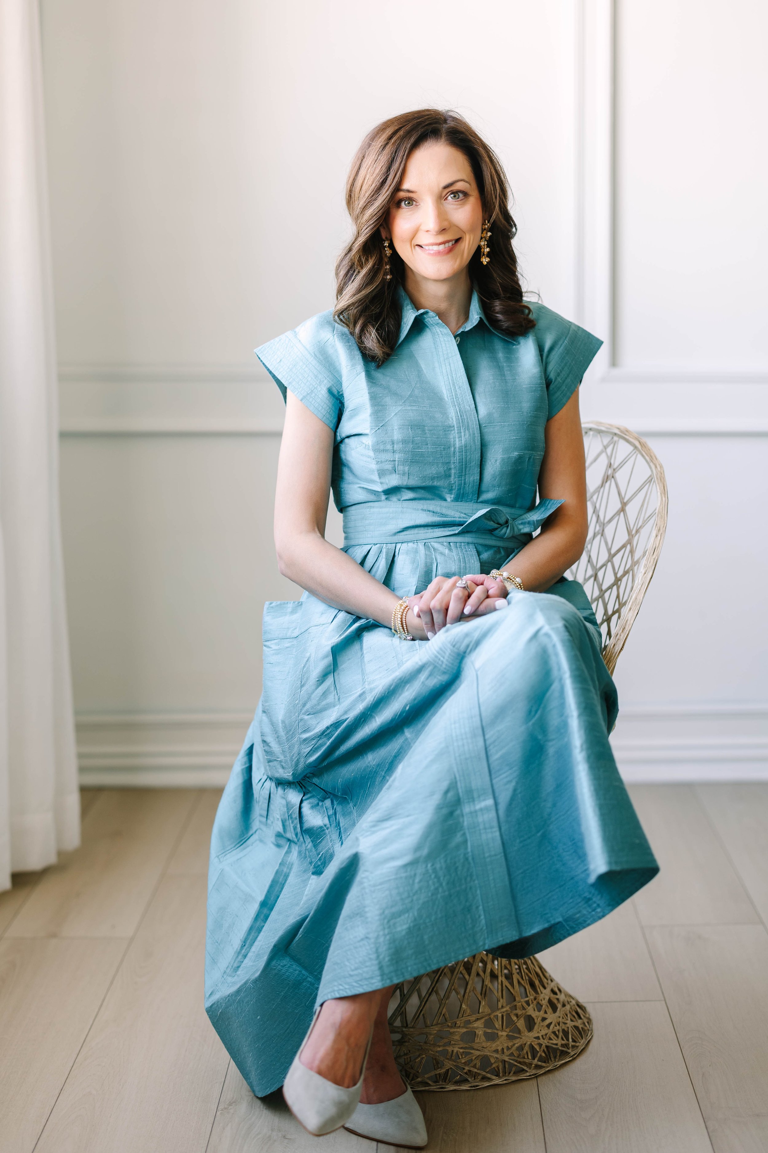 A woman with brown hair and earrings, sitting on a chair in a light blue dress, smiling at the camera in a room with white walls.
