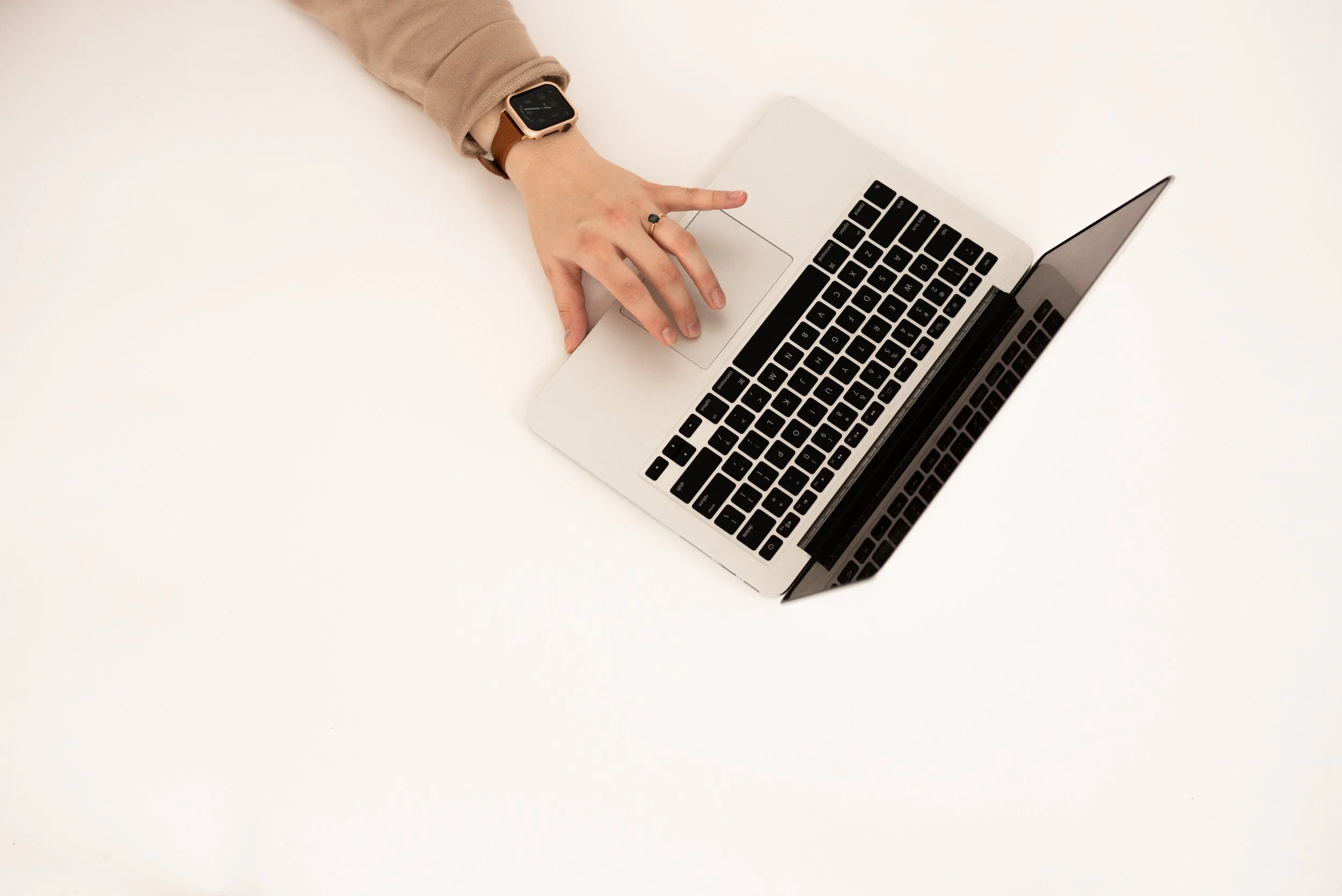 A person's hand with a smartwatch on their wrist, typing on a silver laptop with a black keyboard, on a white surface.
