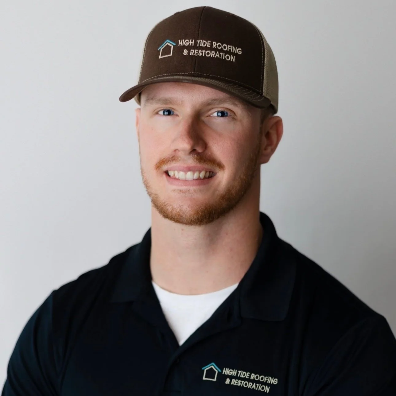 A young man with red hair and a beard, smiling, wearing a black collared shirt and a brown baseball cap with the logo and text 'High Tide Roofing & Restoration'.