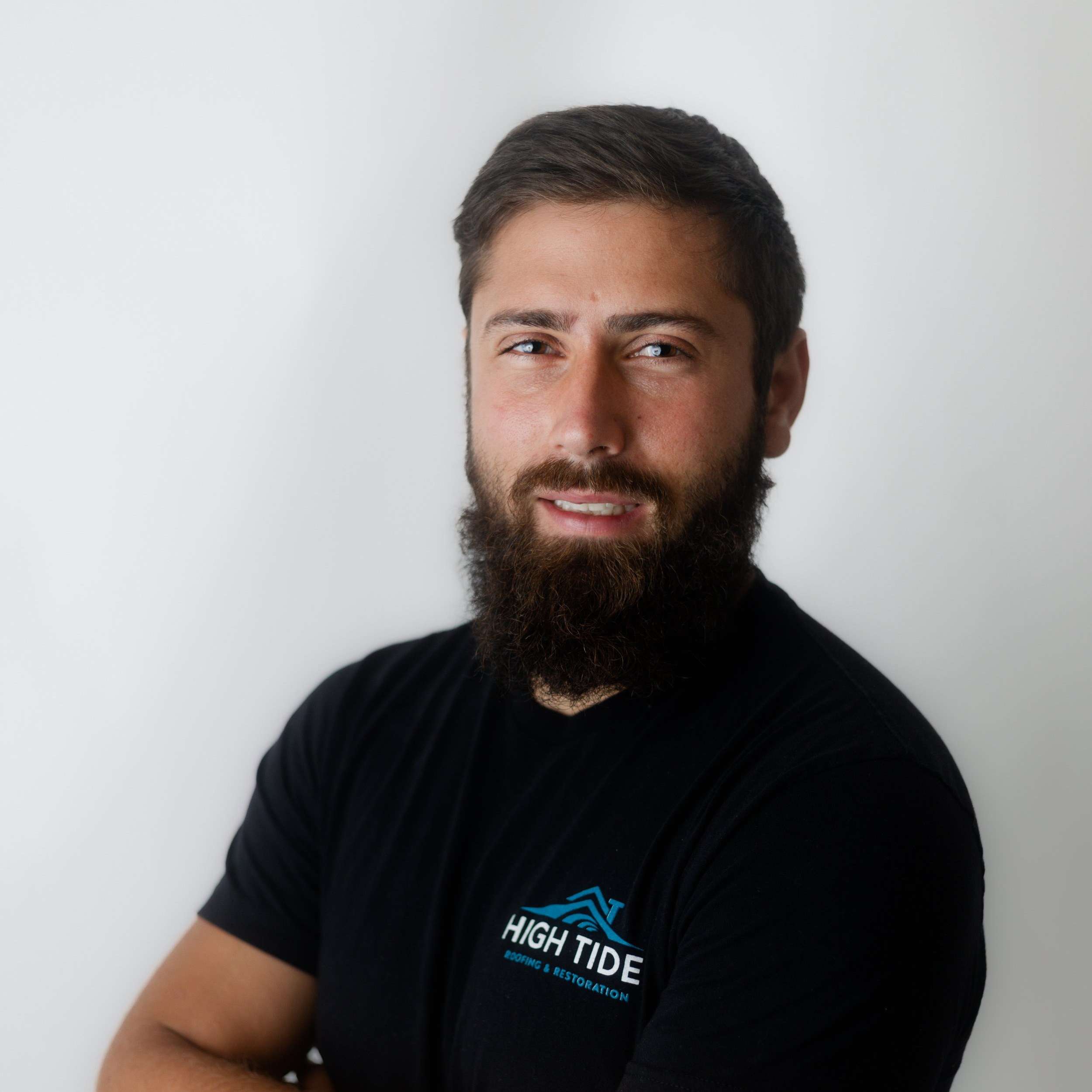 Portrait of a man with a beard and short hair wearing a black T-shirt with 'High Tide' logo, standing against a plain white background.