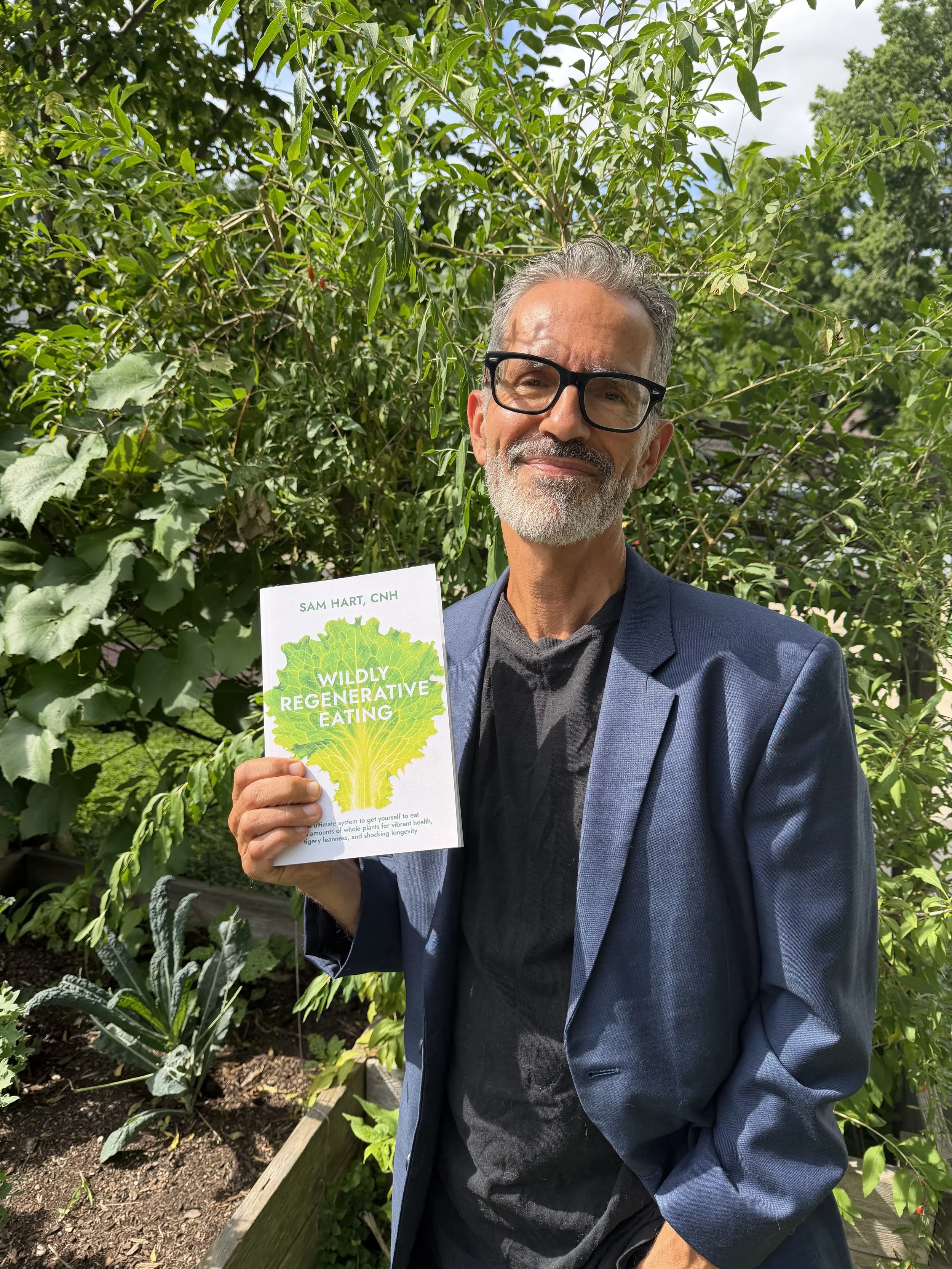 A man with gray hair and beard wearing glasses, a black shirt, and a navy blazer standing in a lush garden. He is holding a book titled 'Wildly Regenerative Eating' by Sam Hart, CNH. The background features various green plants and trees with a partly cloudy sky.