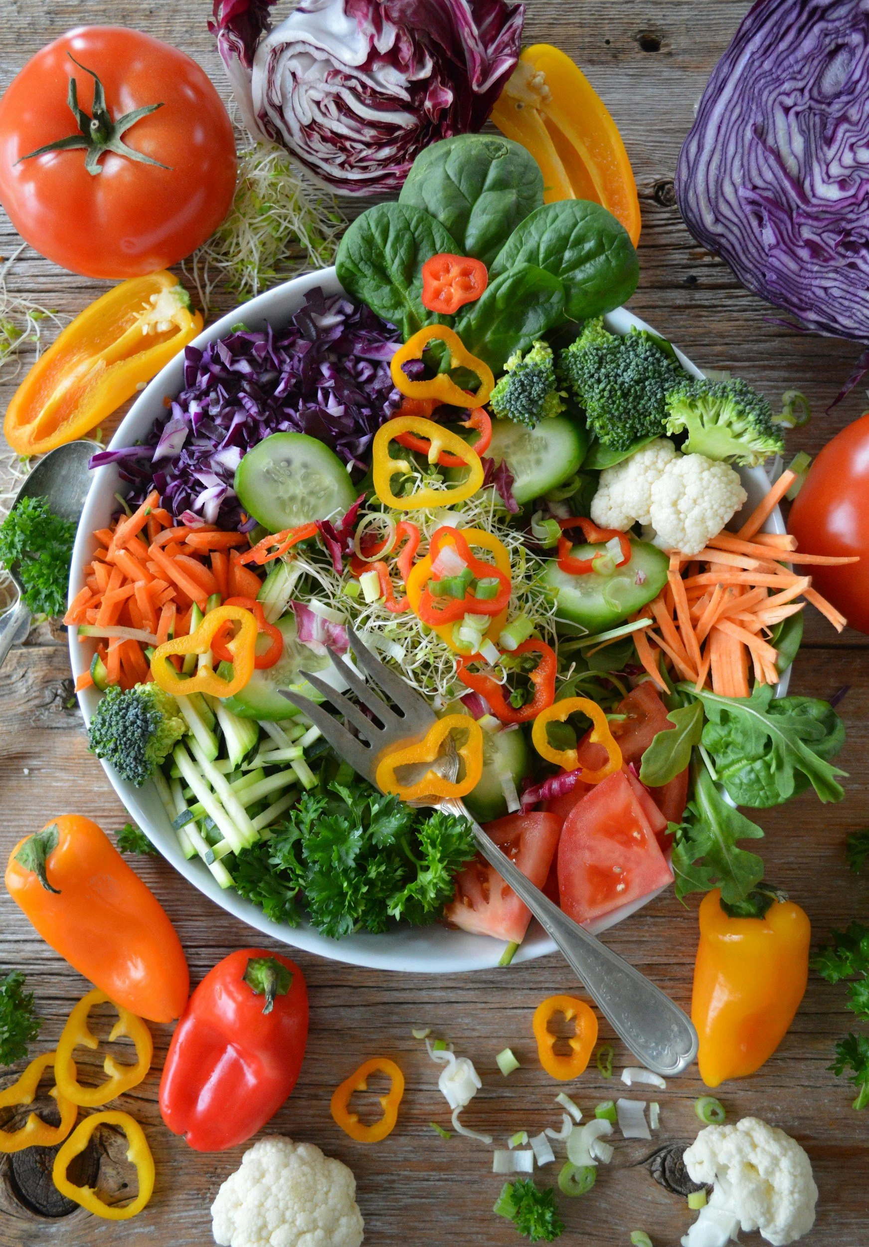 A variety of fresh vegetables arranged on a wooden surface, including tomatoes, yellow and orange bell peppers, cauliflower, broccoli, cucumbers, shredded purple cabbage, shredded carrots, sliced green onions, and fresh herbs like parsley and arugula.