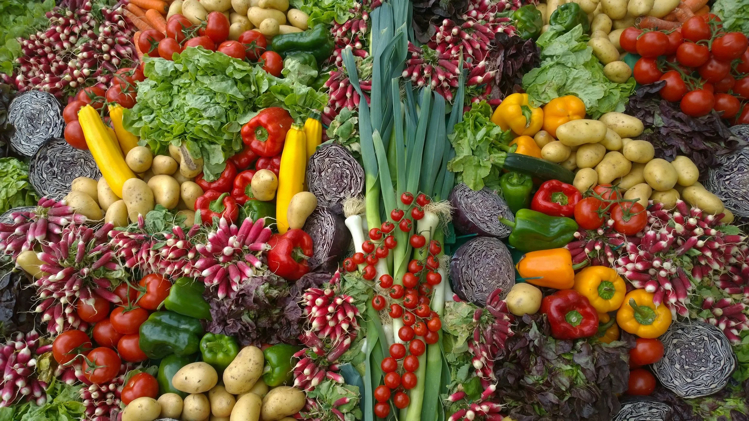 A colorful assortment of fresh vegetables including cherry tomatoes, potatoes, bell peppers, radishes, zucchinis, lettuce, and purple cabbage.