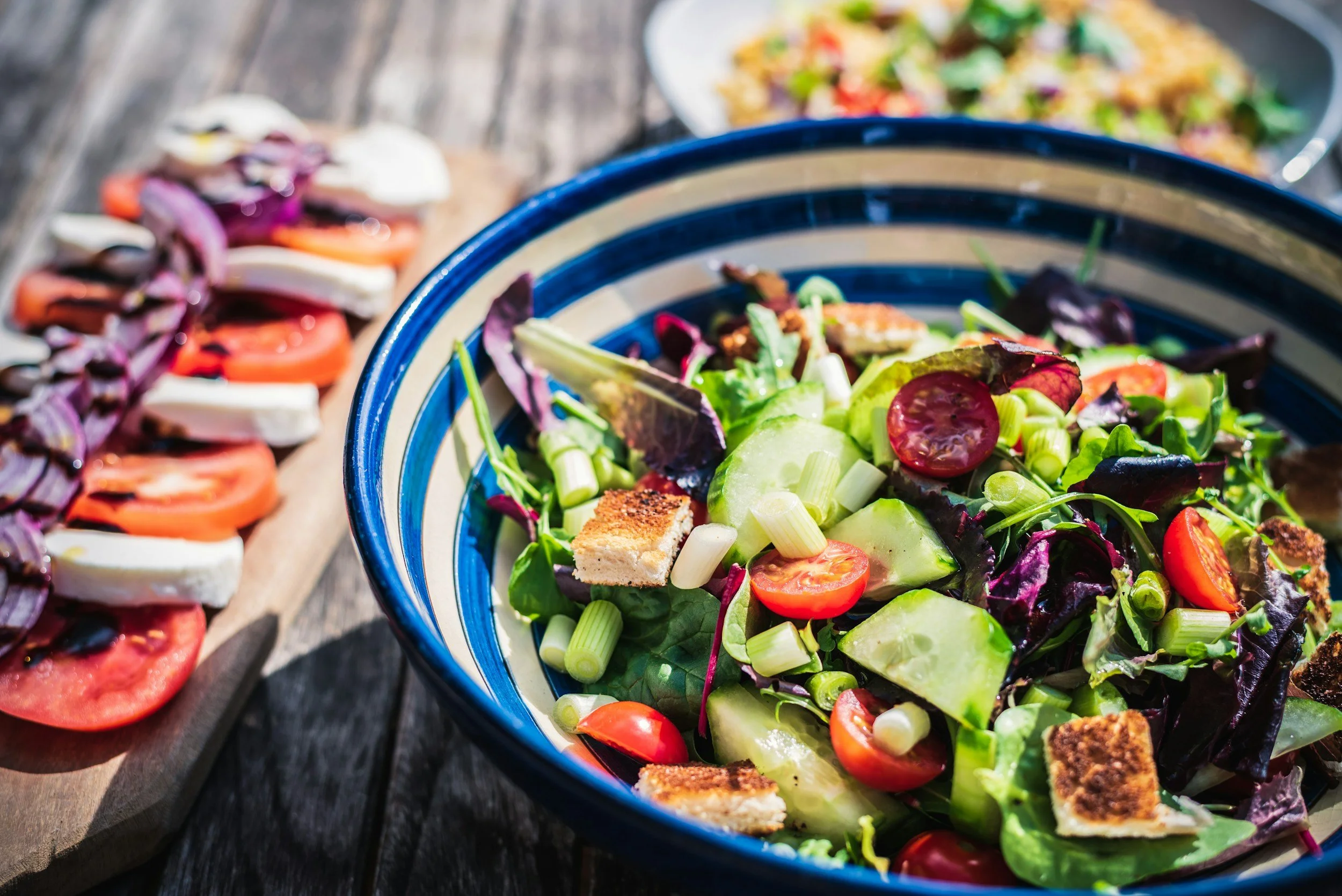 A colorful salad with cherry tomatoes, cucumber, red onion, mixed greens, and croutons in a blue and beige striped bowl, set on a wooden table.