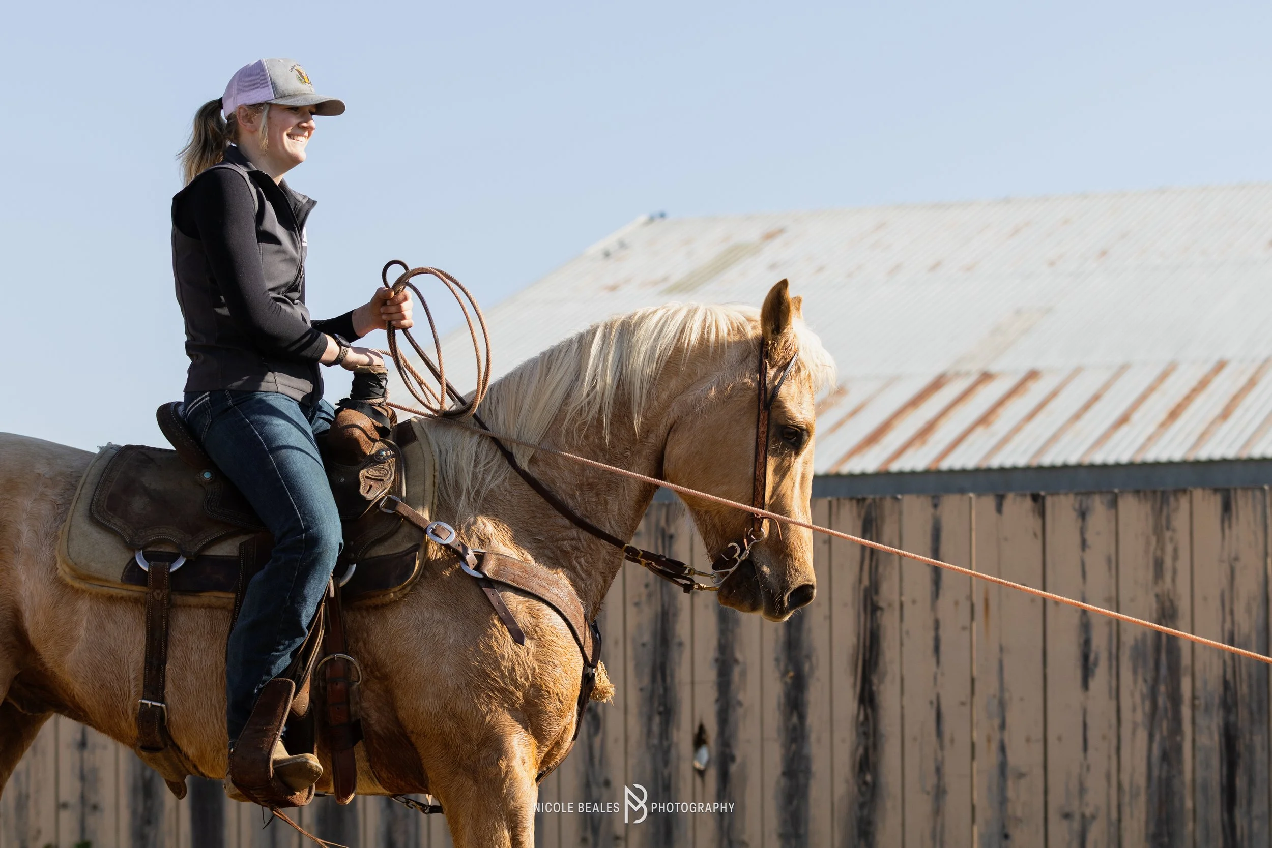 A woman riding a light brown horse with a cream mane, holding reins, outdoors with a wooden barn and clear blue sky in the background.