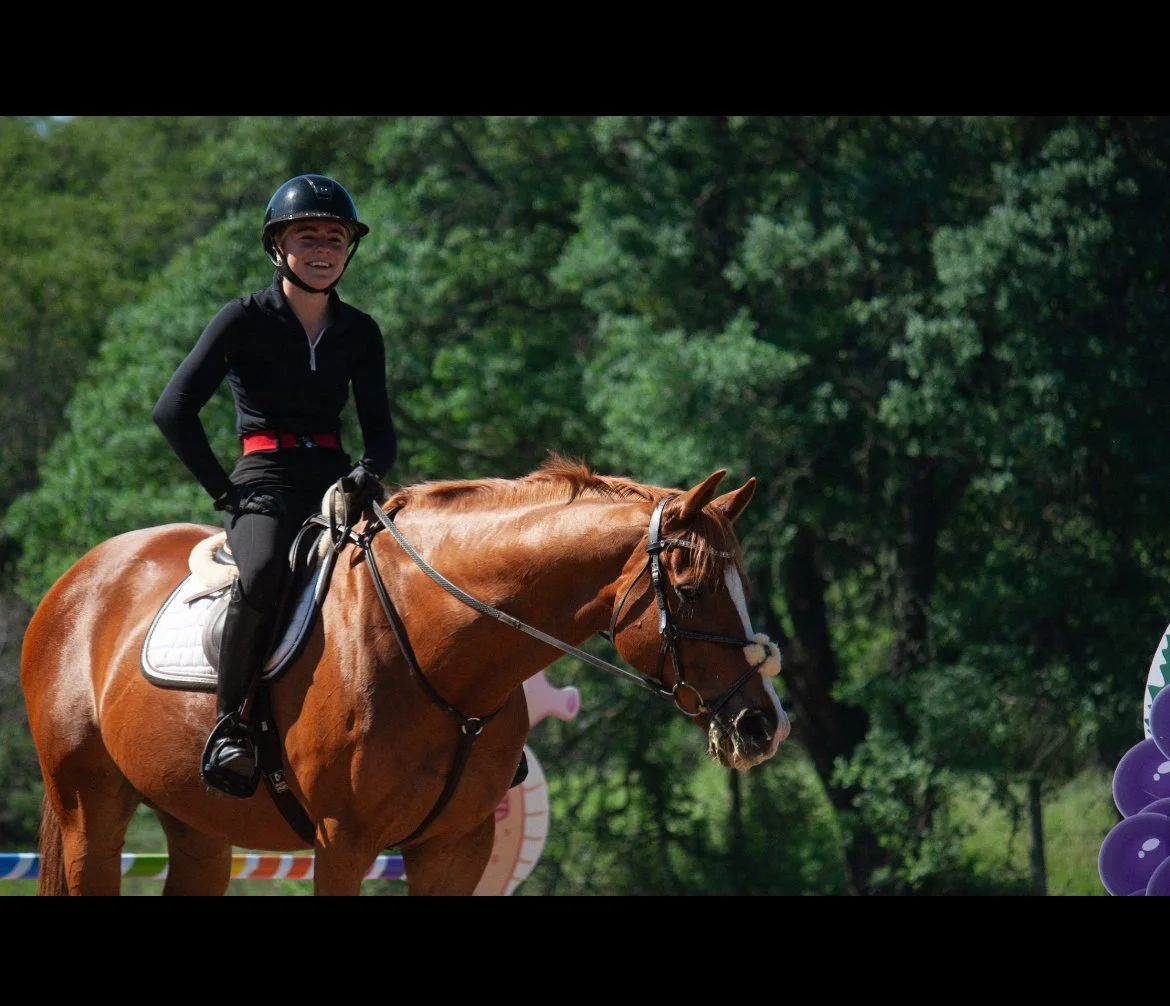A young woman wearing black riding gear and a helmet rides a chestnut horse with a white blaze on its face in an outdoor setting with green trees and purple balloons.