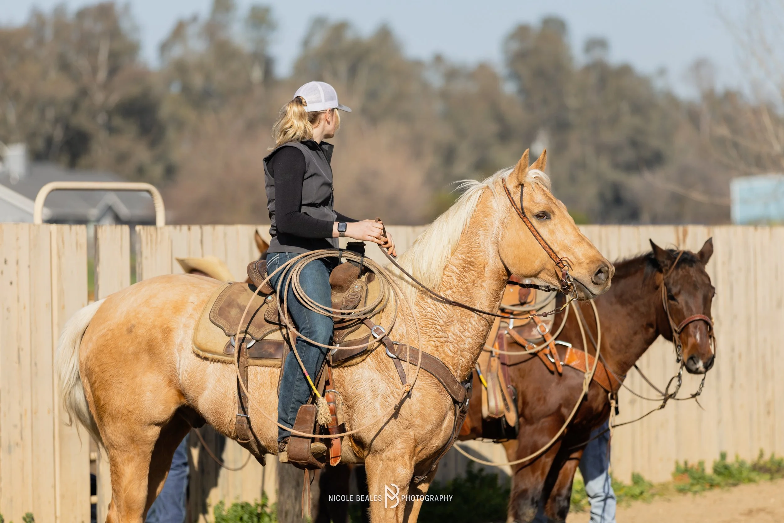 A woman riding a tan horse with a blonde mane, wearing a black vest, jeans, and a white cap, holding the reins. There is another dark brown horse in the background, and a wooden fence surrounds the area.