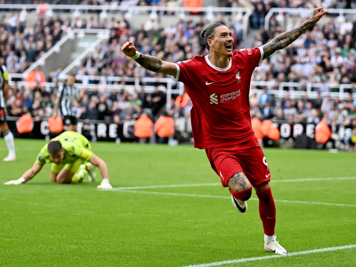 Soccer player in red uniform celebrating a goal on the field, with opposition goalkeeper in green kneeling on the ground and blurred spectators in the background.