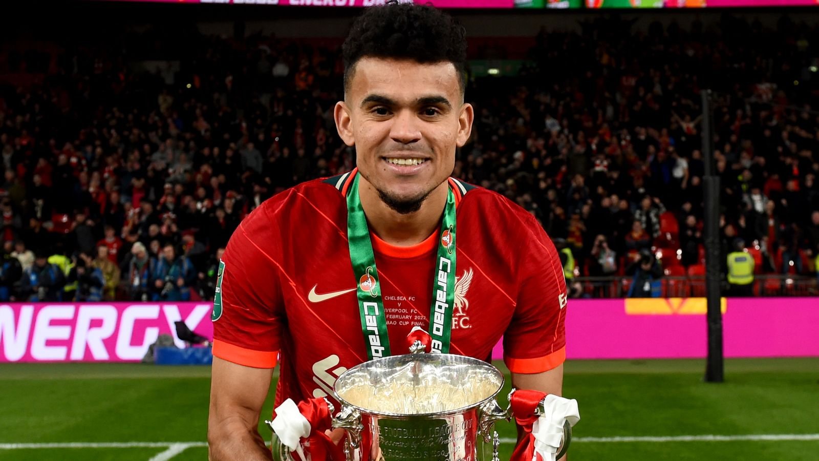 Soccer player in red jersey holding trophy with green ribbon, celebrating victory in stadium.