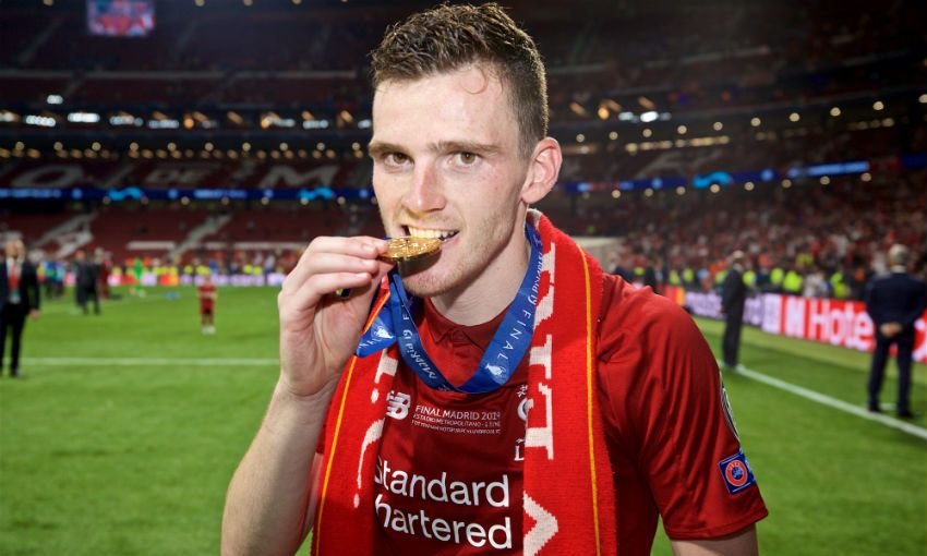 Soccer player wearing a red jersey and a Champions League medal, biting the medal on a stadium field with fans in the background.