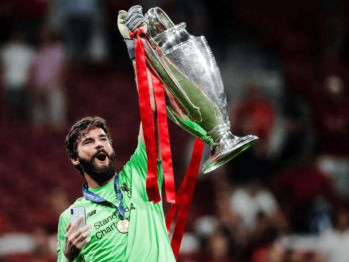 A soccer player wearing a green goalkeeper jersey celebrates holding a large trophy with red ribbons on a soccer field, surrounded by a blurred crowd in the background. The player has a medal around his neck and a phone in his hand.