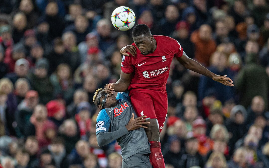 Two soccer players jumping for a header during a match, with one wearing a red jersey and the other a blue one. They are competing for control of the ball in front of a crowd.