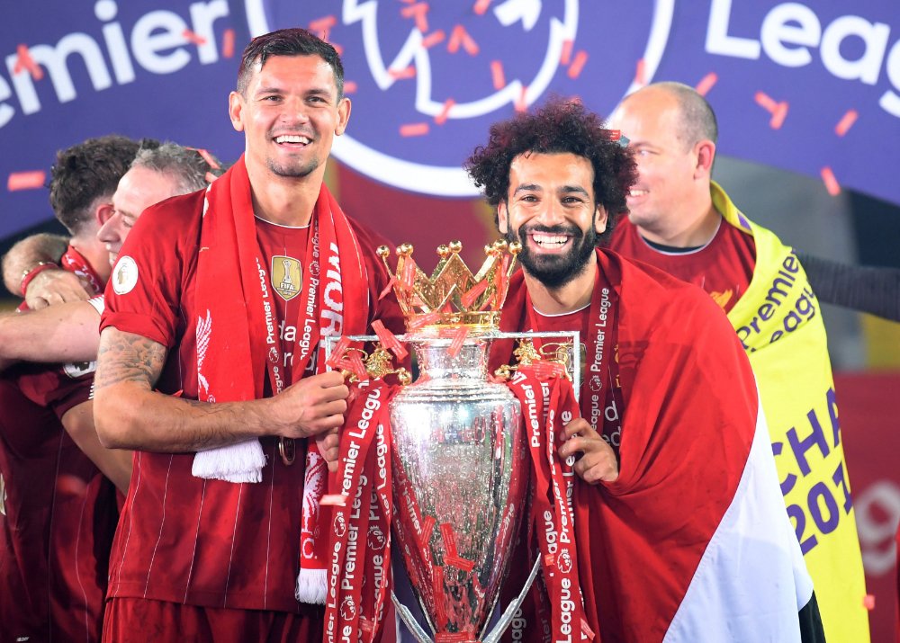 Soccer players celebrating with the Premier League trophy, wearing red jerseys and scarves, in front of a banner with the Premier League logo.