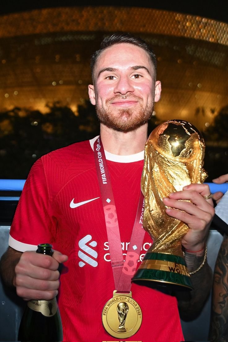 Soccer player in a red jersey holding the FIFA World Cup trophy and a champagne bottle, wearing a winner's medal.