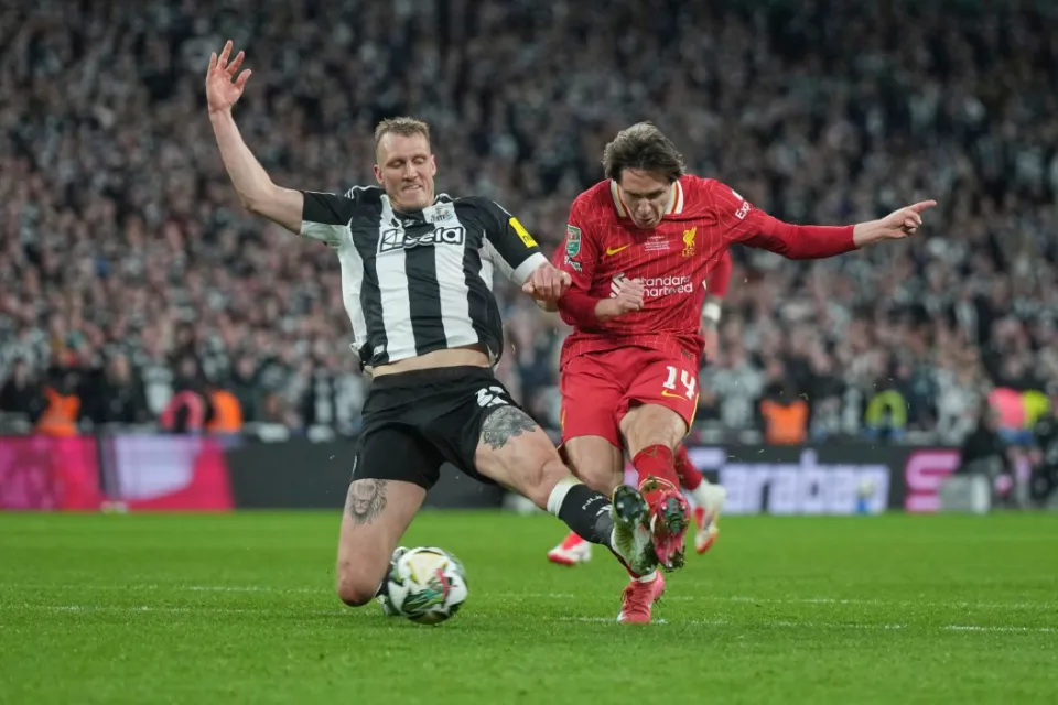 Soccer match action with players from opposing teams in red and black-and-white jerseys, competing for the ball on the field with a blurred crowd in the background.